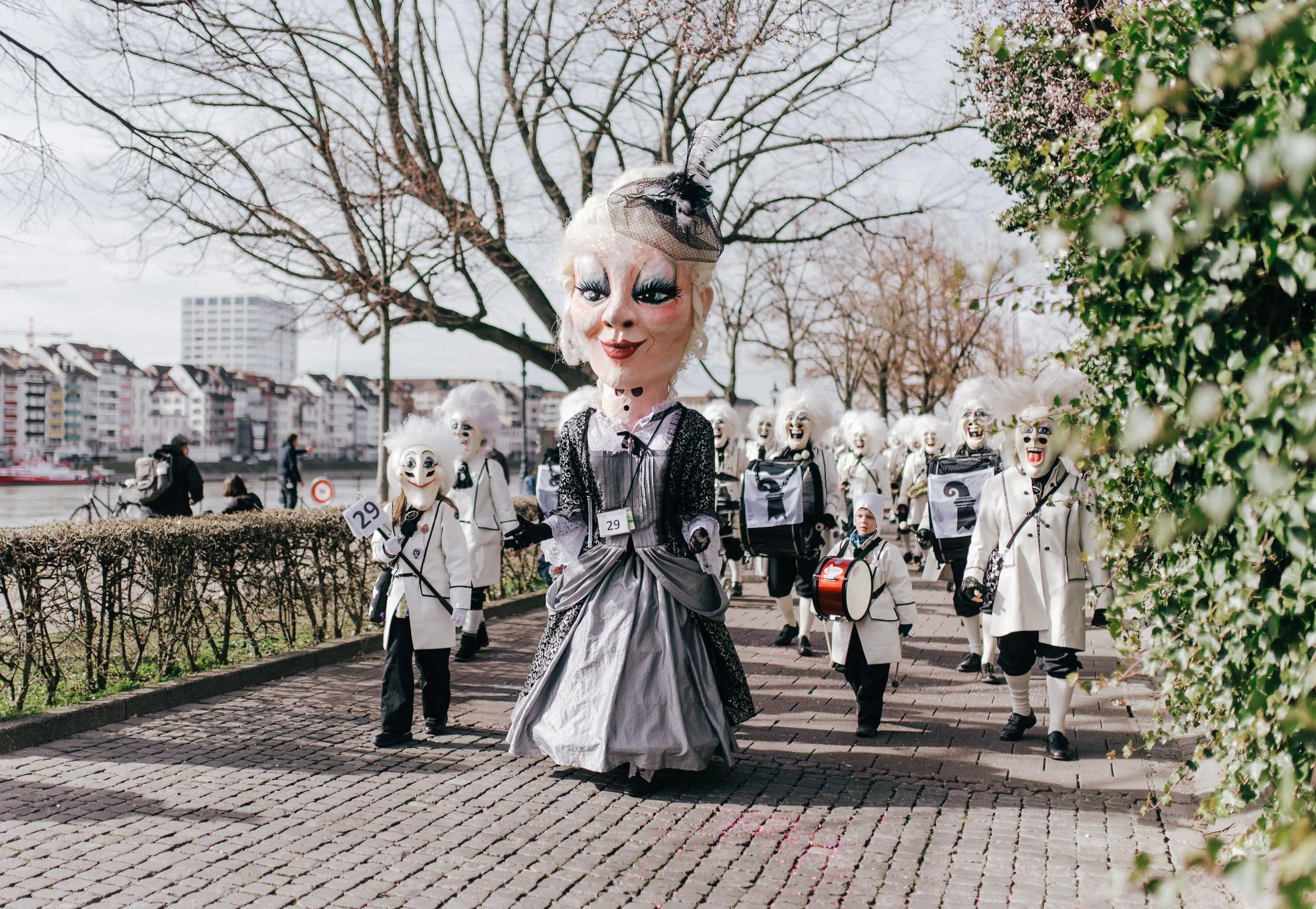 Gruppe von Menschen in Karnevals- oder Faschingskostümen mit großen Masken und auffälligem Make-up, die an einem Spaziergang entlang eines Flusses in einer europäischen Stadt teilnehmen.