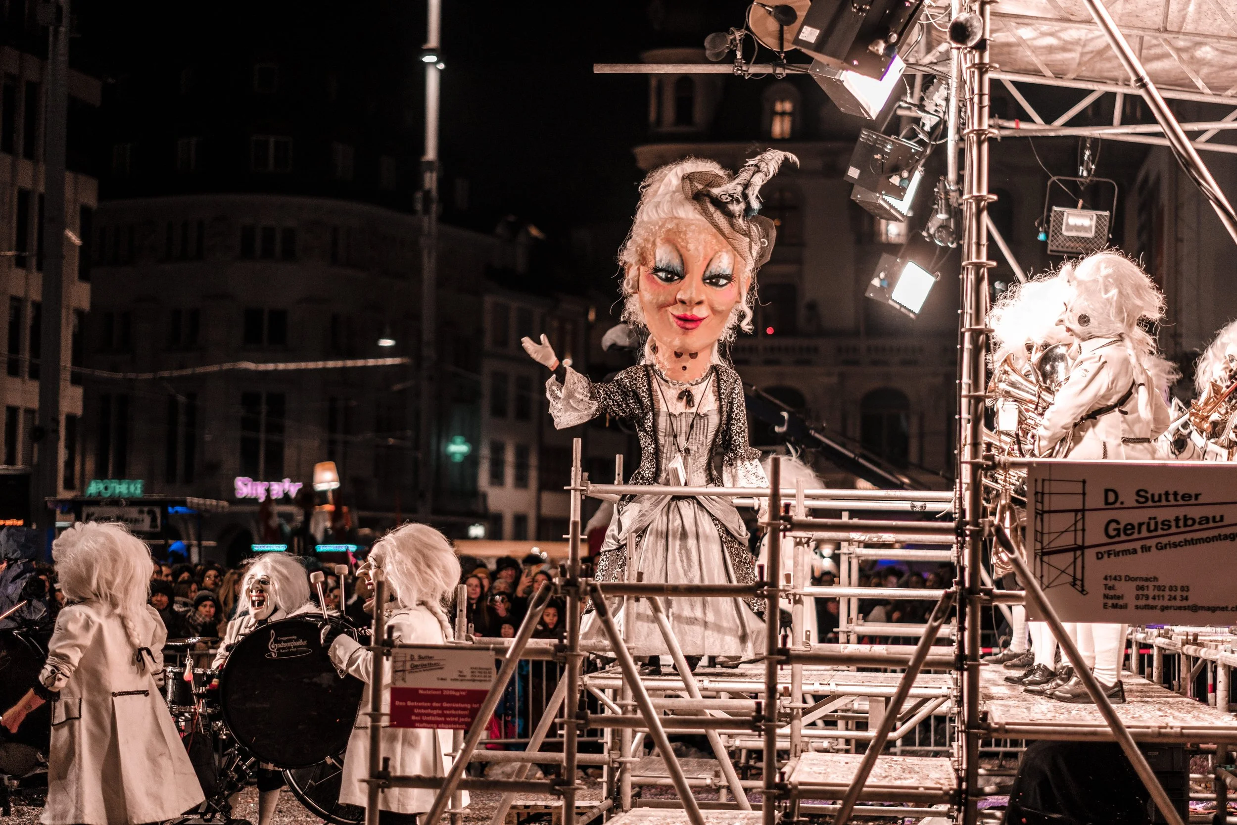 Maskierte Figuren in einem Festwagen bei einer nächtlichen Parade, mit großen Puppen und einer Menschenmenge im Hintergrund, beleuchtet mit Straßenlaternen.