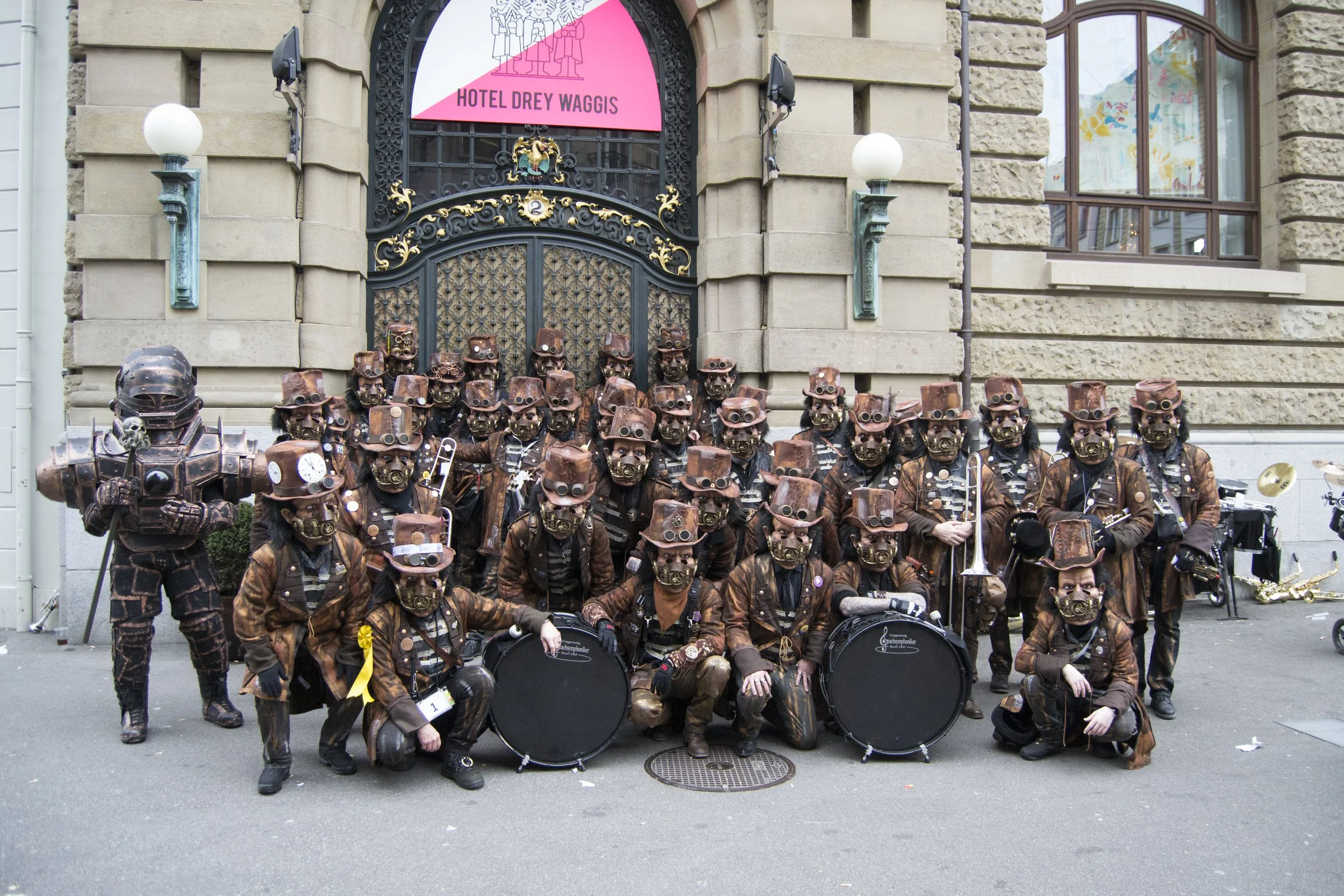 Eine Gruppe von Menschen in Steampunk-Kostümen und Masken, die Musikinstrumente halten, steht vor einem historischen Gebäude mit einer Metalltür und einem pinken Schild mit der Aufschrift 'Hotel Drey Waggis.'