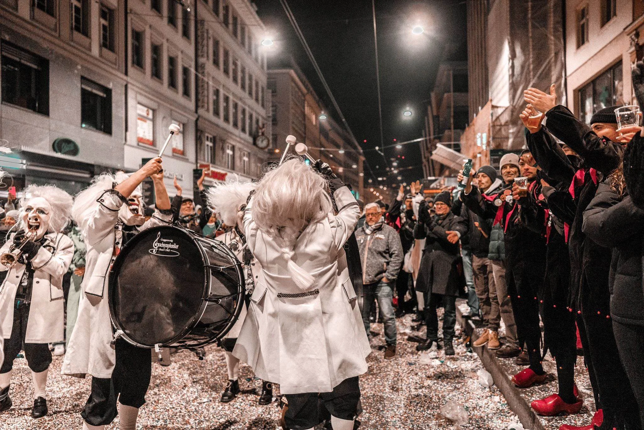 Menschen in Karnevals- oder Faschkostümen, die auf einer Straßenszene feiern, Pokal und Bonbons werfend, mit confetti auf dem Boden, nachts in einer Stadt.