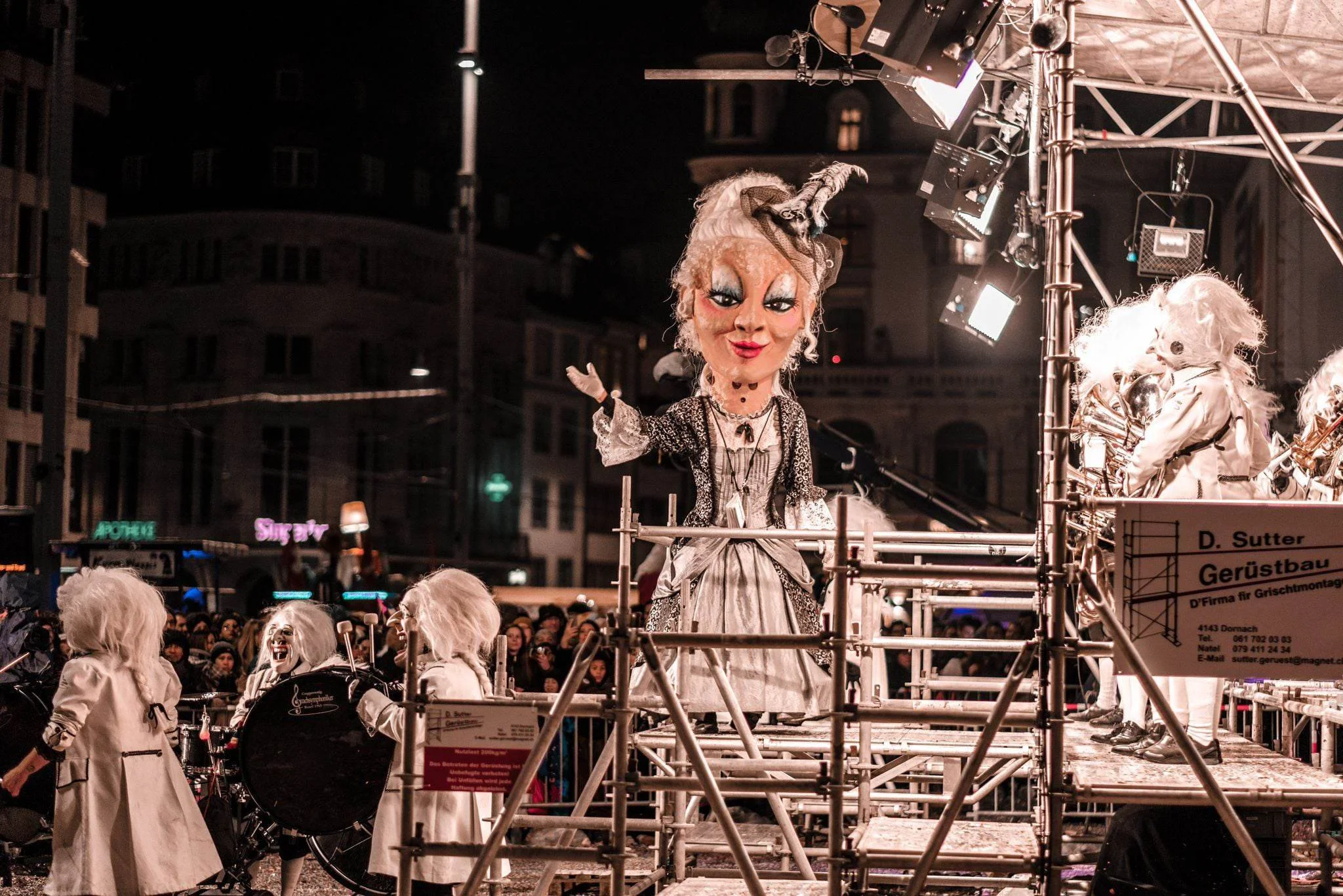 Großes, bizar geschminktes Marionetten-Kunstwerk auf einem Fahrzeug bei einer nächtlichen Parade, umgeben von kostümierten Personen und einem Publikum.