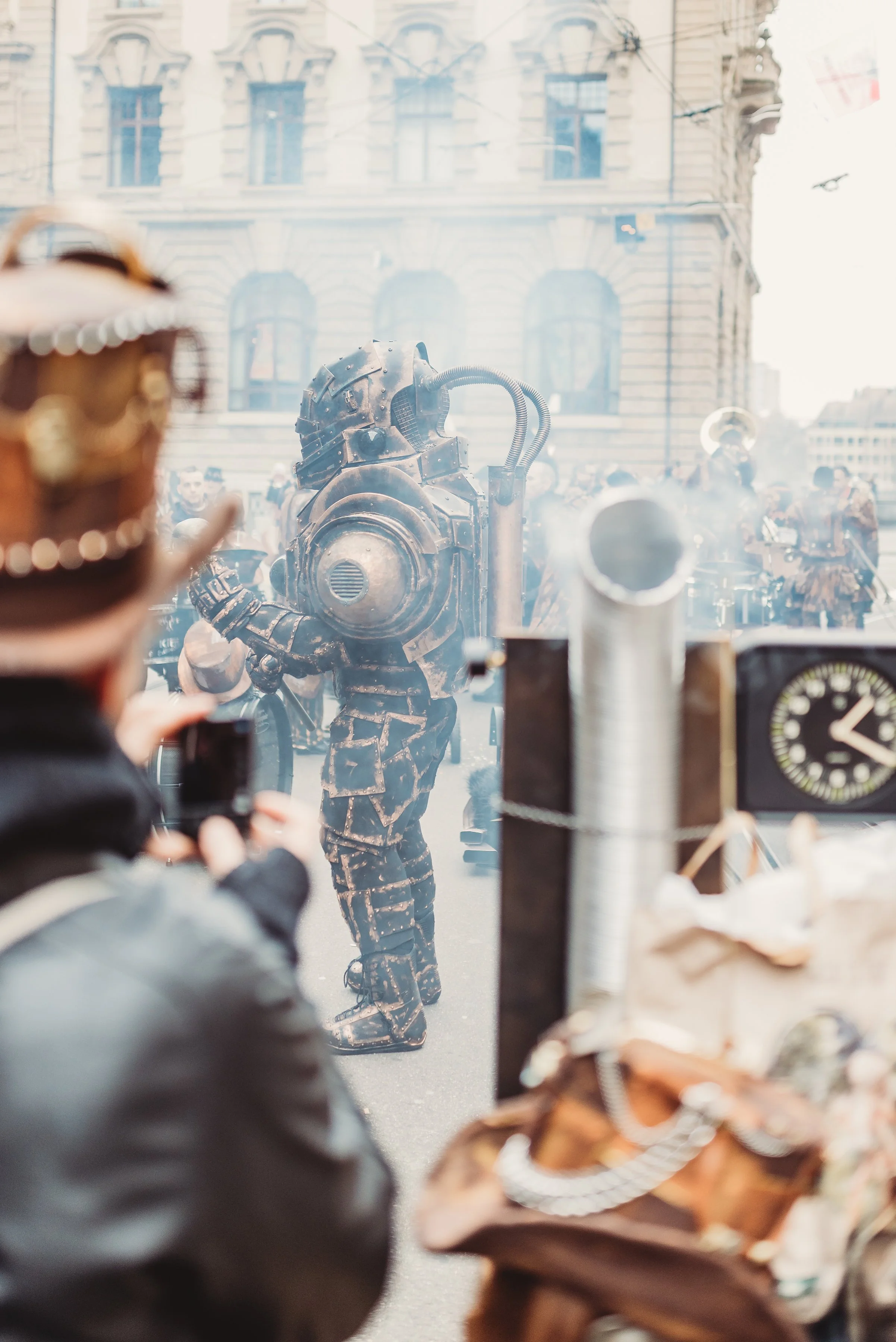Person beobachtet einen Mann in einem futuristischen Roboterkostüm durch ein Fenster. Im Hintergrund sind weitere Menschen und Gebäude zu sehen.