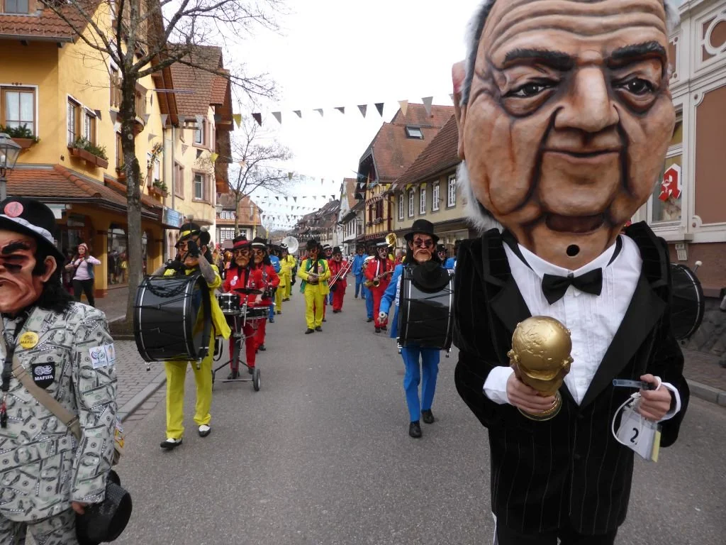 Gruppe von Menschen in Karnevalskostümen mit großen Masken, die in einer Straße mit bunten Gebäuden und festlicher Dekoration marschieren.