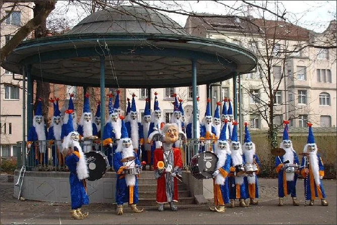 Eine Gruppe von Menschen in Nikolauskostümen spielt auf einer kleinen Parkbühne, die mit blauen Jacken, weißen Bärten und roten Mützen gekleidet sind, während ein clownartiger Geselle in der Mitte steht.