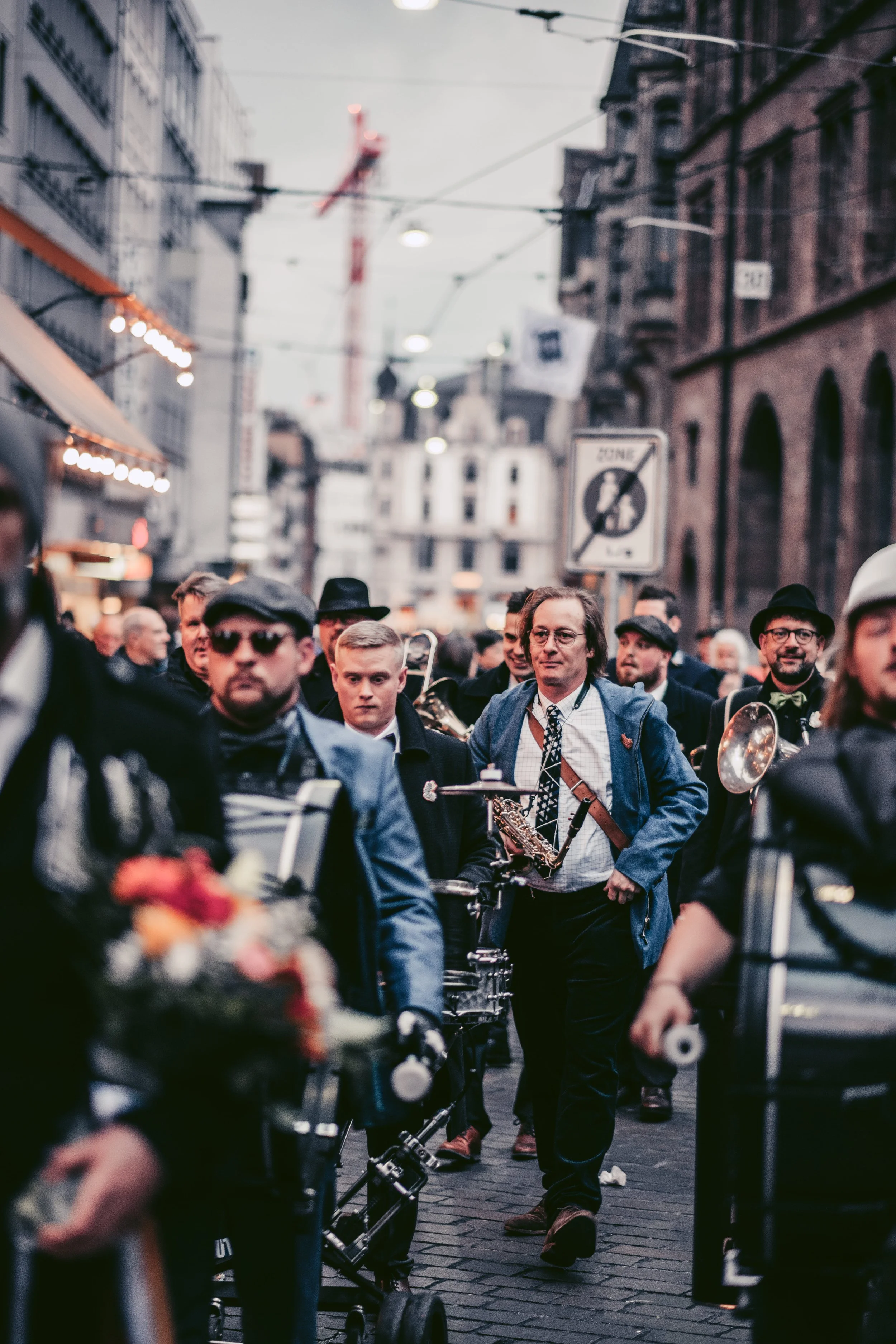Menschen schlendern durch eine belebte Straße, einige tragen Hüte und Sonnenbrillen, im Hintergrund ein Kran und historische Gebäude.