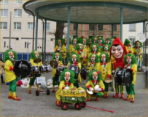 Eine Gruppe von Clowns in bunten Kostümen vor einer alten Parkbühne, viele mit einem großen Lächeln, einige spielen Trommeln und haben eine Laterne, es wirkt wie eine Zirkus- oder Karnevalsauftritt.