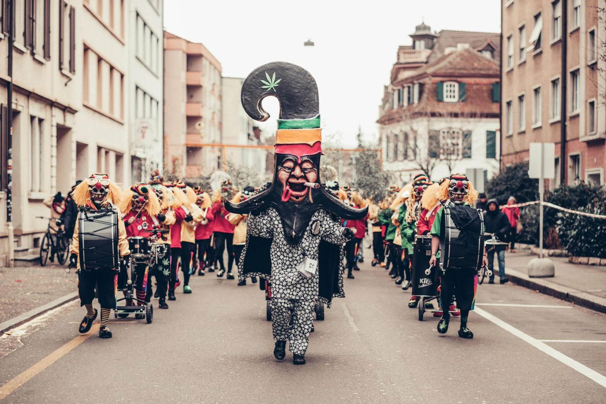 Menschen in farbenfrohem Karnevalskostüm, einige im Clown- und Maskenkostüm, marschieren auf einer Straße in einer Stadt, mit alten Gebäuden im Hintergrund, bei einem offiziellen Karnevalsumzug.