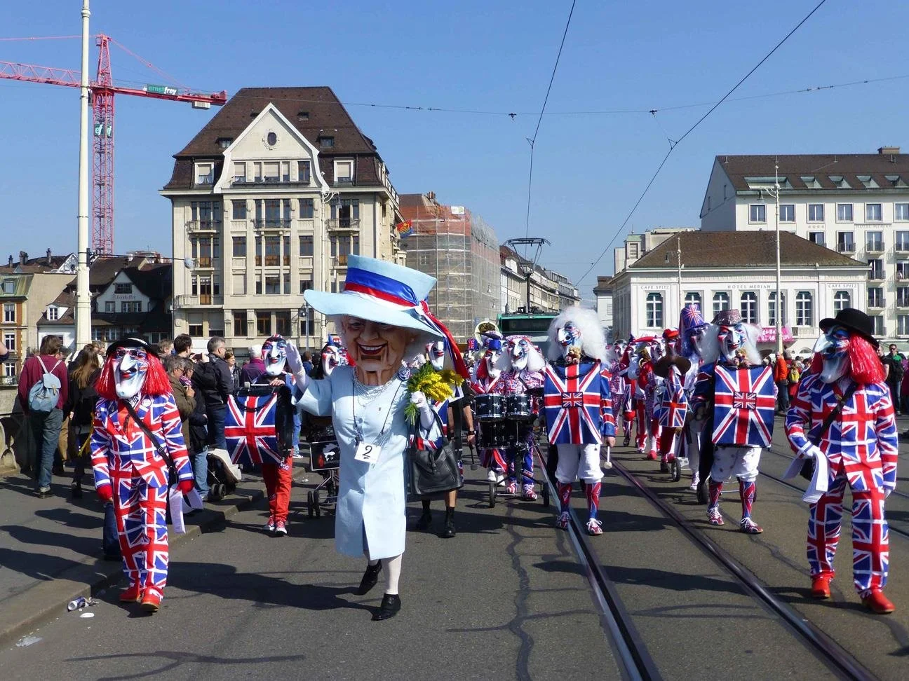 Menschen in einer Parade tragen Kostüme mit der britischen Flagge und Masken, eine Person trägt einen großen Hut und einen weißen Mantel, im Hintergrund befinden sich Gebäude und eine Baustelle.