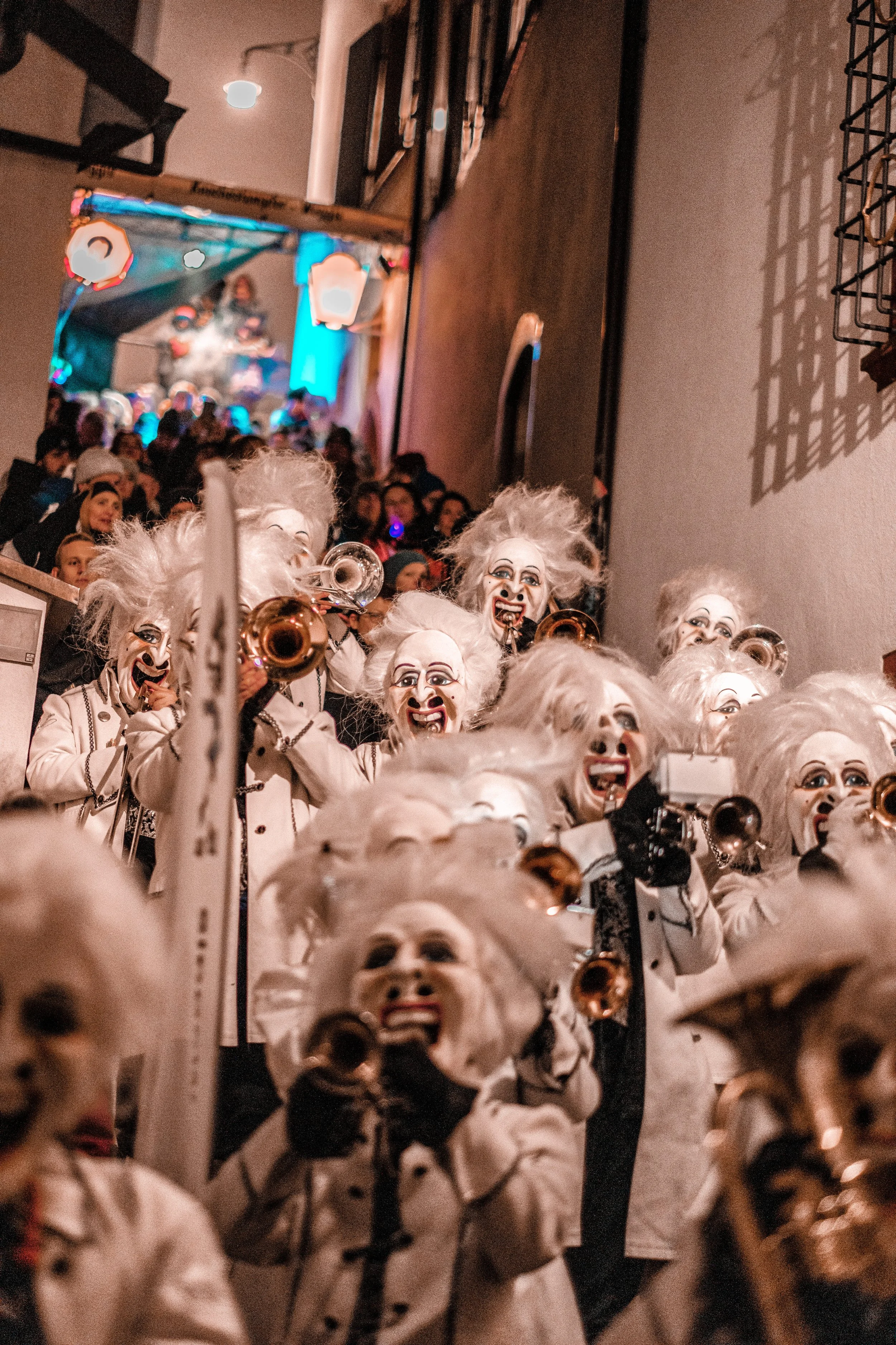 Gruppe von Menschen in Karnevalskostümen mit weißen Perücken und Masken, die Musikinstrumente spielen, im Hintergrund eine Menschenmenge bei einer Feier.