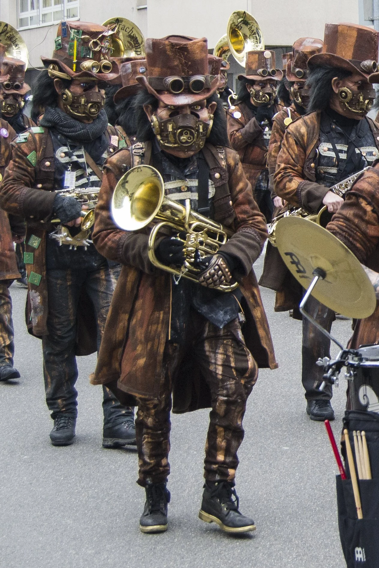 Menschen in Kostümen im Steampunk-Stil bei einer Parade, tragen braune Zylinder, Masken mit mechanischen Elementen, und spielen Musikinstrumente wie Trompete und Schlagzeug.