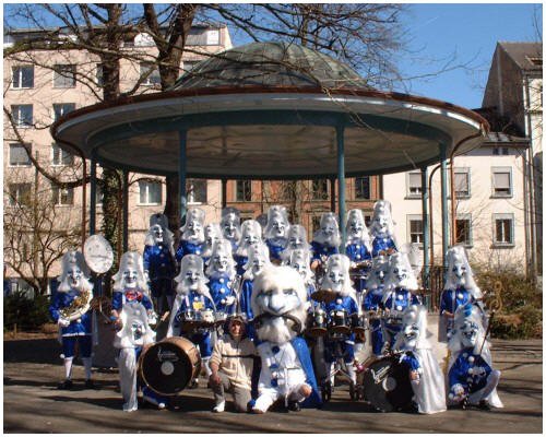Gruppe von Menschen in Karnevalskostümen mit weißen Masken, blauen Kostümen und Musikinstrumenten vor einem Riesenrad im Freien, umgeben von Bäumen und Gebäuden.