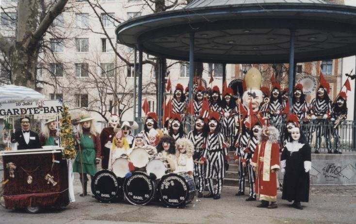 Verschiedene Menschen in Clowns- und Zirkuskostümen, die auf einem Karussell und vor einem Gebäude mit Bäumen im Hintergrund stehen. Es scheint eine festliche Veranstaltung zu sein.