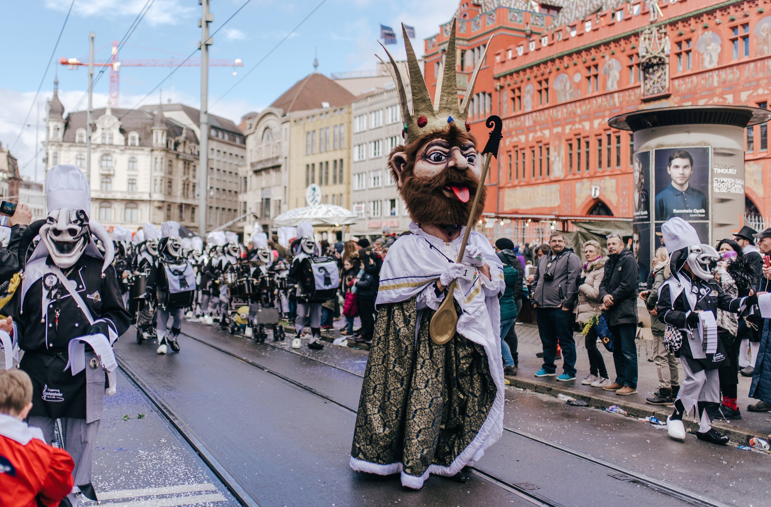 Menschen in Karnevalskostümen und Masken, eine große Figur mit einem Zepter, Parade in einer Stadt mit historischen Gebäuden im Hintergrund.