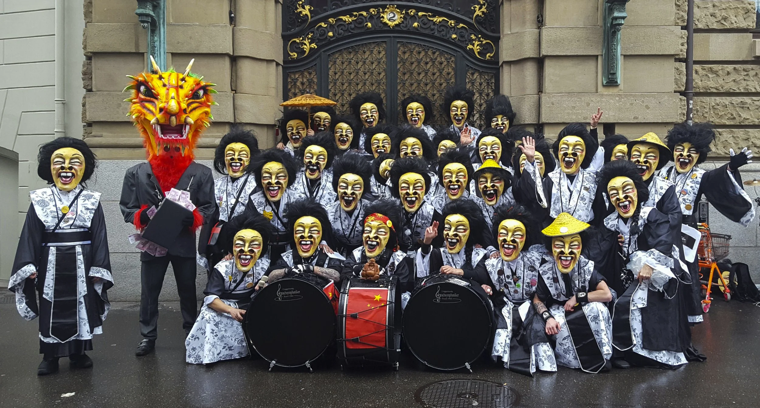 Gruppe von Menschen in traditionellen japanischen Kostümen mit Masken, einem riesigen Drachenkopf, Trommeln, vor einem historischen Gebäude.