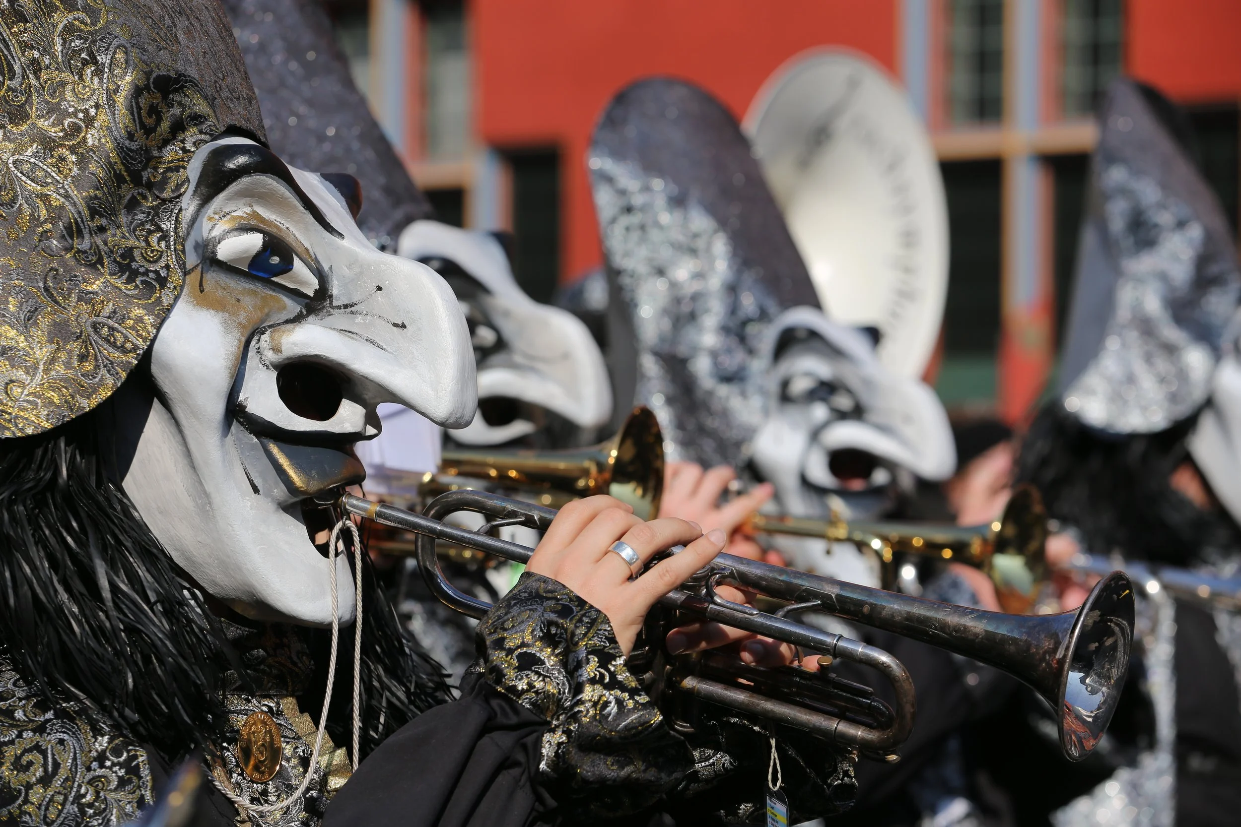 Musiker in Karnevals- oder Faschenkostümen mit weißen Masken, die auf einem Musikfestival 'Motto' spielen, während sie Trompeten blasen.