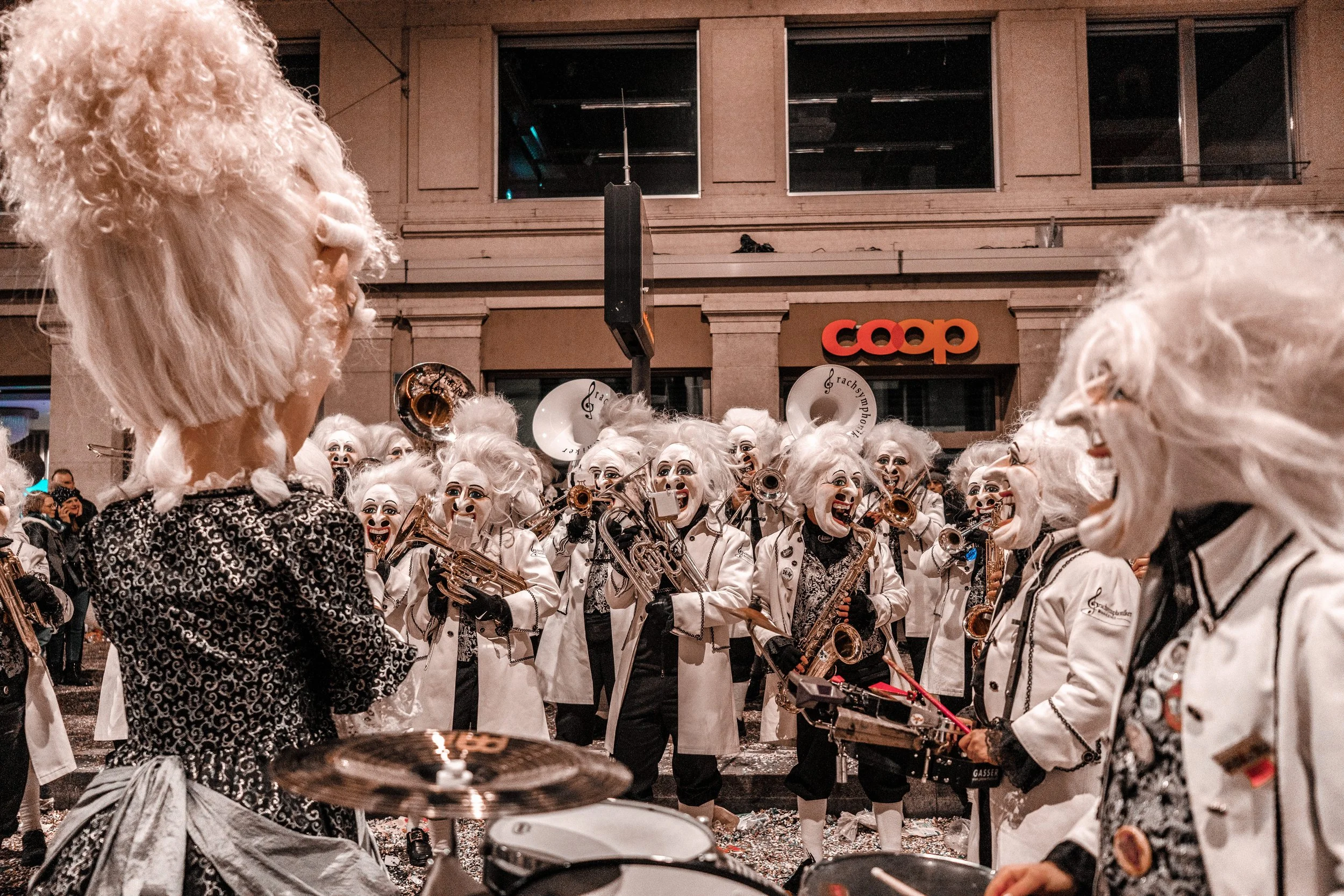 Eine Gruppe von Menschen in Kostümen mit weißen Perücken und bemalten Gesichtern spielen Musikinstrumente während einer Straßenparade, im Hintergrund ein Gebäude mit einem Coop-Schild.