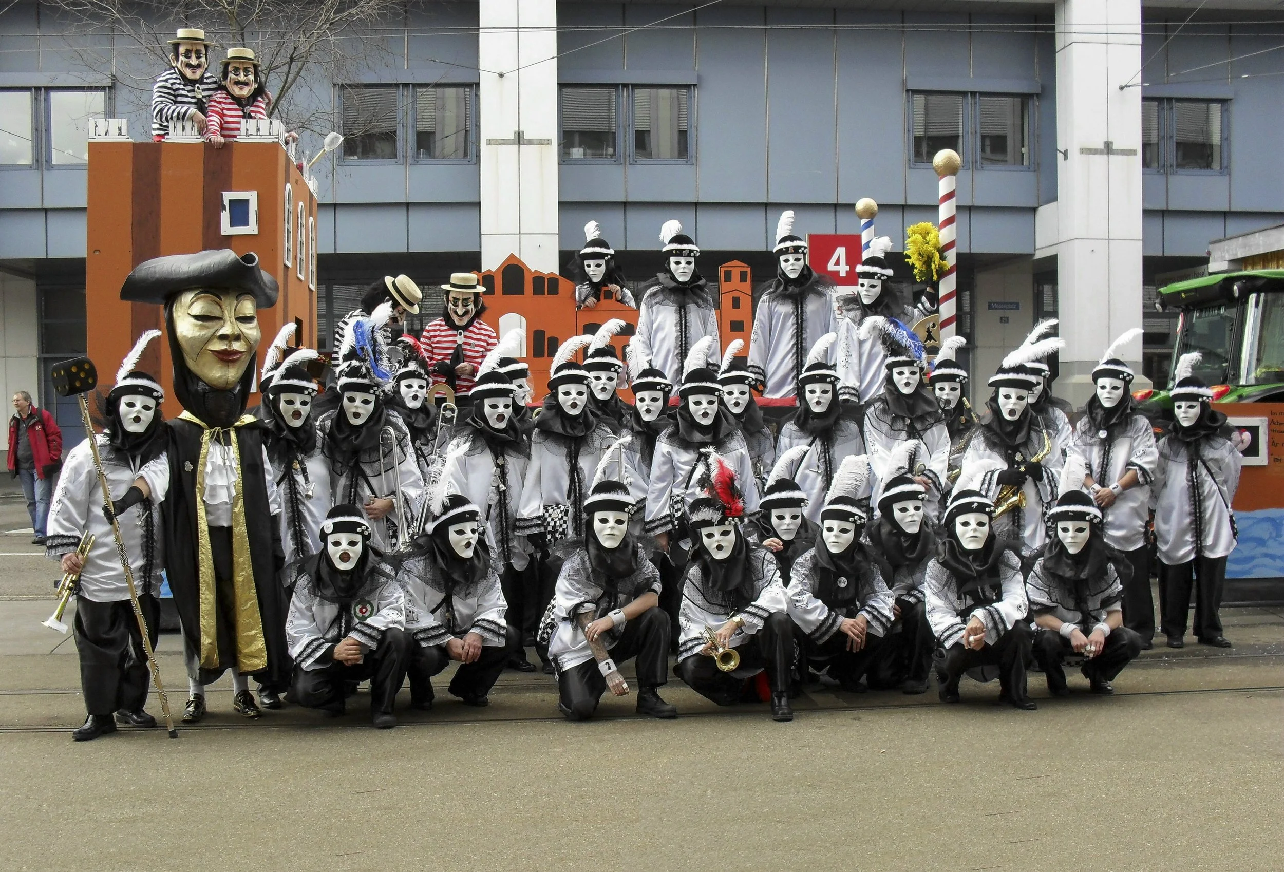 Gruppe von Menschen in Karnevals- oder Faschingskostümen, die weißen Masken, schwarze und weiße Kleidung, einige mit Federn und Schreibmaschinen tragen, bei einer Parade oder Feier in einer Stadt.