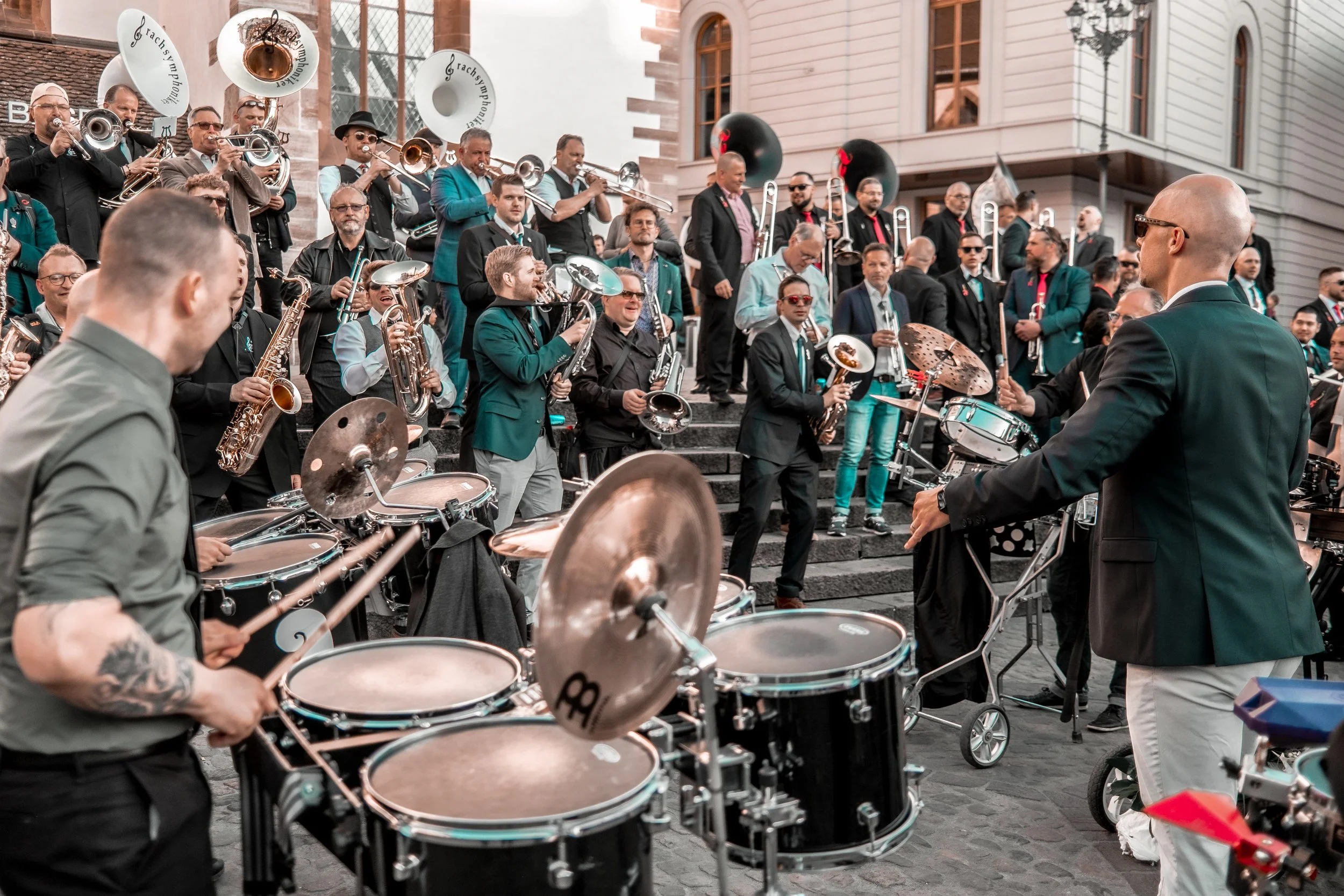 Eine große Gruppe von Musikern, hauptsächlich mit Blechblasinstrumenten und Saxophonen, die auf der Treppe vor einem historischen Gebäude performen. Vorne erkennt man einen Dirigenten, der das Orchester leitet, während im Hintergrund weitere Musiker 