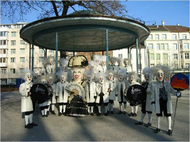 Eine Gruppe von Menschen in Clowns-Kostümen mit weißen Pelz-Masken und verschiedenen Musikinstrumenten steht auf einem Platz in einer Stadt, im Hintergrund sind Wohnhäuser sichtbar.