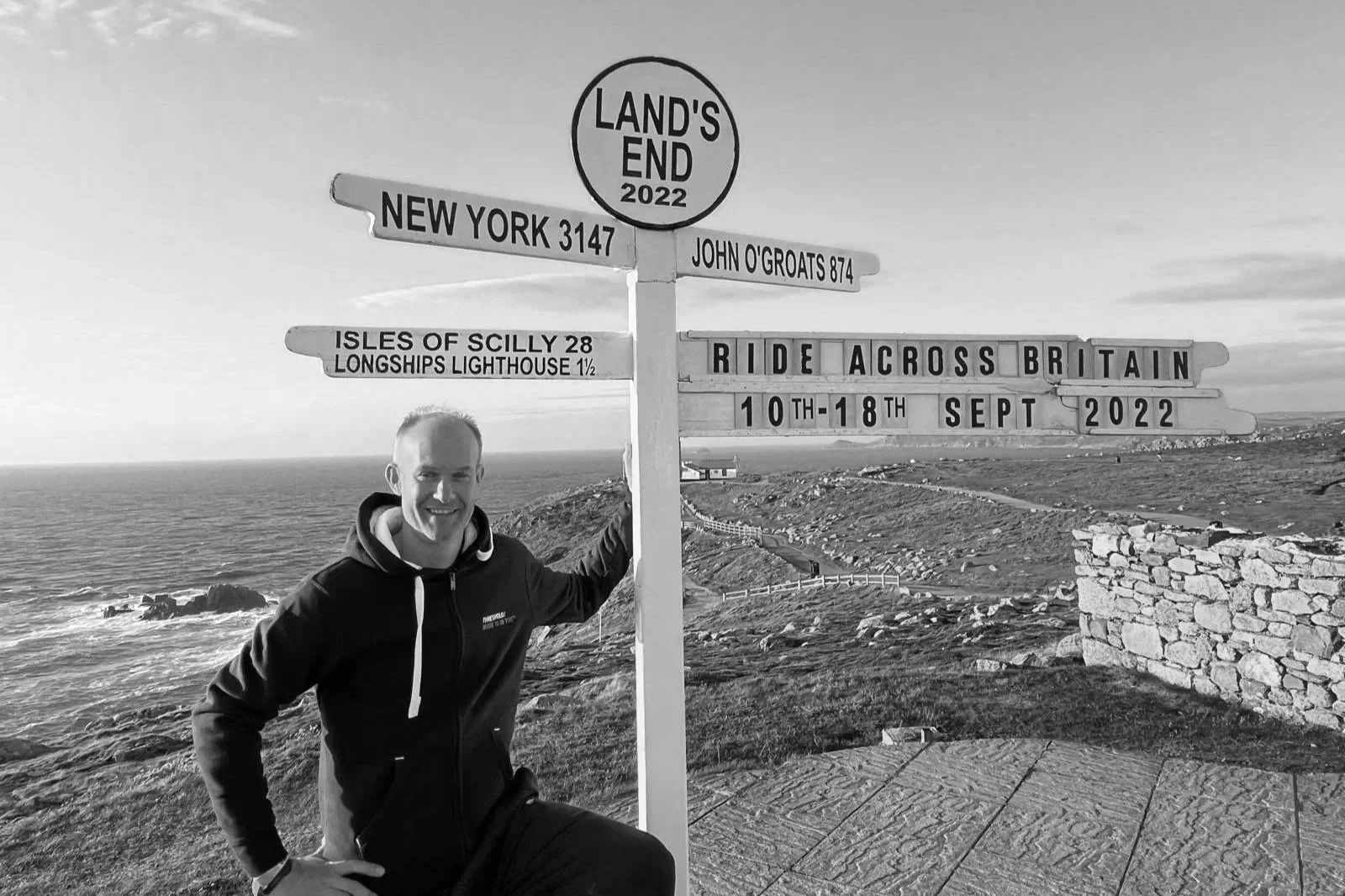 A man smiling and sitting near a signpost at Land's End, England, in 2022. The sign indicates distances to locations including New York, Isles of Scilly, and John O'Groats, with a message to ride across Britain from September 10th to 18th, 2022, with a scenic coastal background.