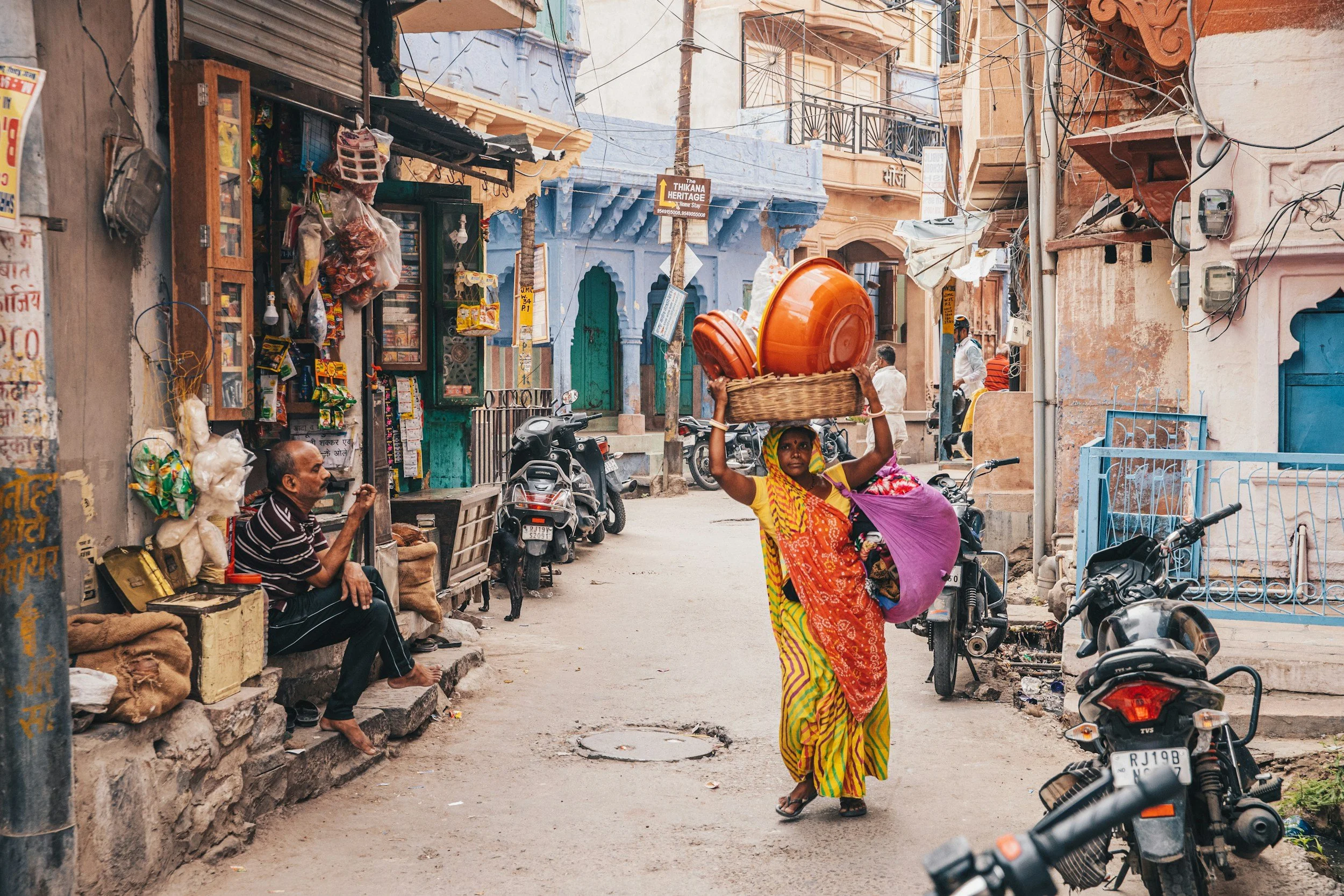 A woman in a colorful sari carries a basket with orange bowls on her head as she walks down a busy street in India, with motorcycles parked along the side and shops and people in the background.