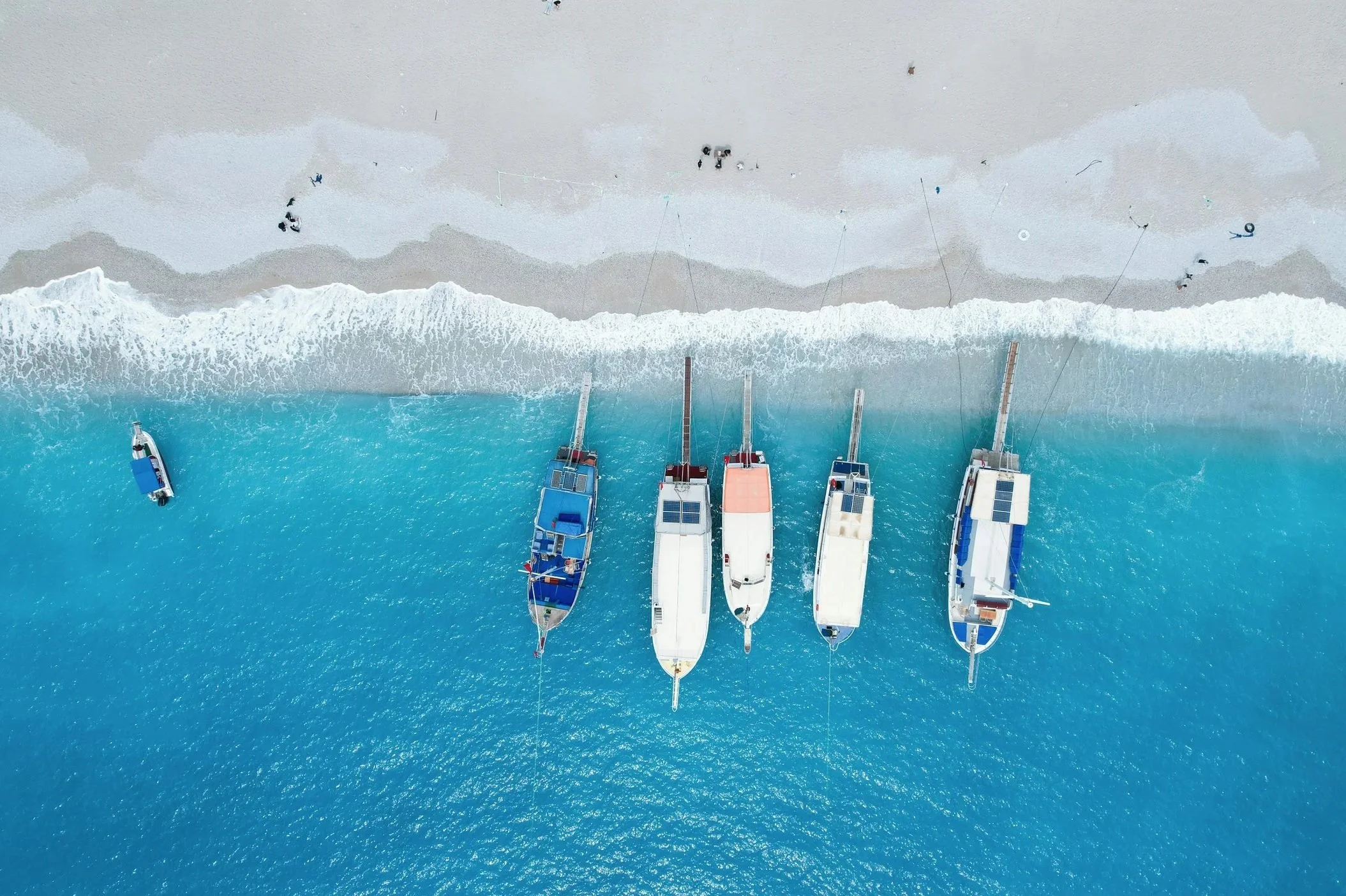 An aerial view of five boats anchored in turquoise water near a sandy beach, with a few people walking along the shoreline and some fishing from the beach.
