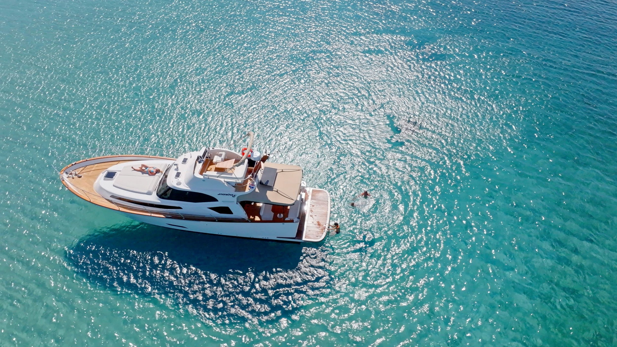 A white yacht floating on clear turquoise water with a person relaxing on the deck and two people swimming nearby.
