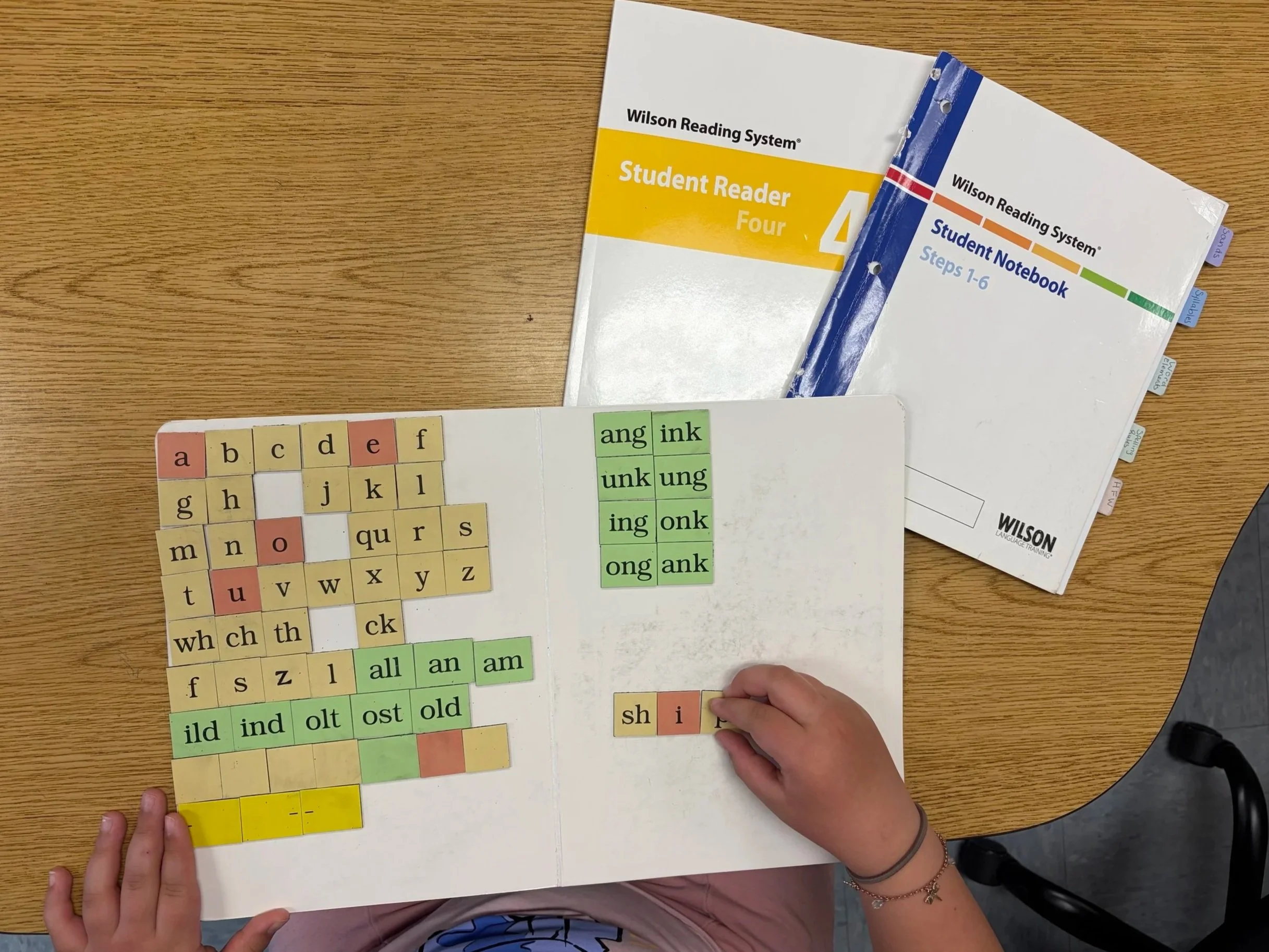 A person holding a sorting activity with letter tiles and word parts on a bulletin board, school books on a wooden table. Wilson Reading System dyslexia tutoring.