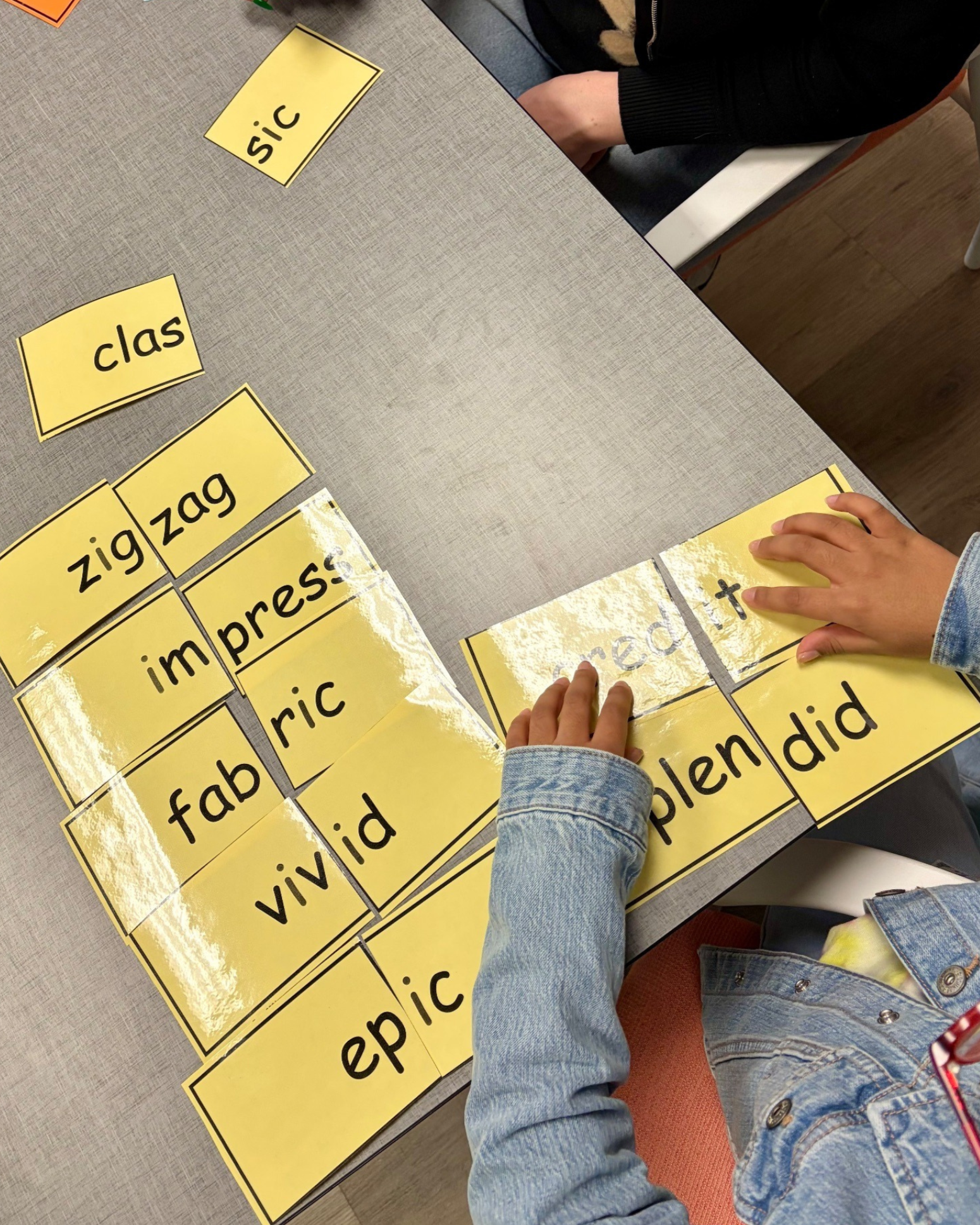 Child wearing blue denim jacket and sitting at a table with yellow word cards, rearranging words such as 'class,' 'zebra,' 'vivid,' and 'happy'. Literacy tutoring session.