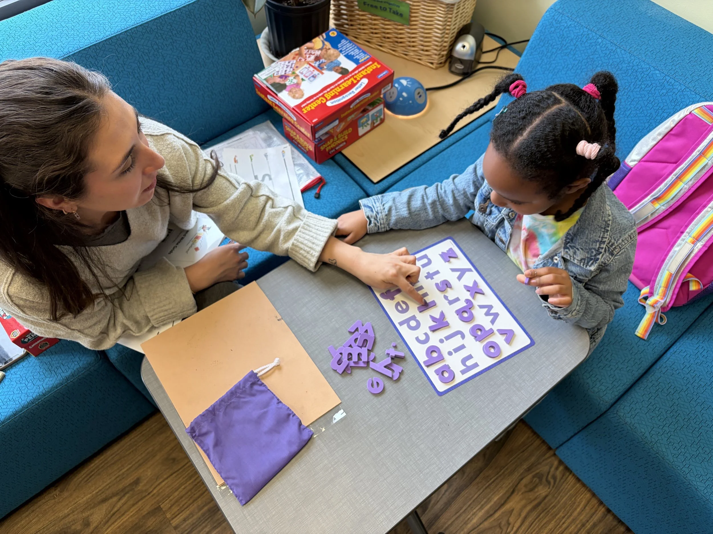 Two young girls are sitting at a table working on educational activities involving purple foam letters and a laminated alphabet worksheet. Tutoring session.