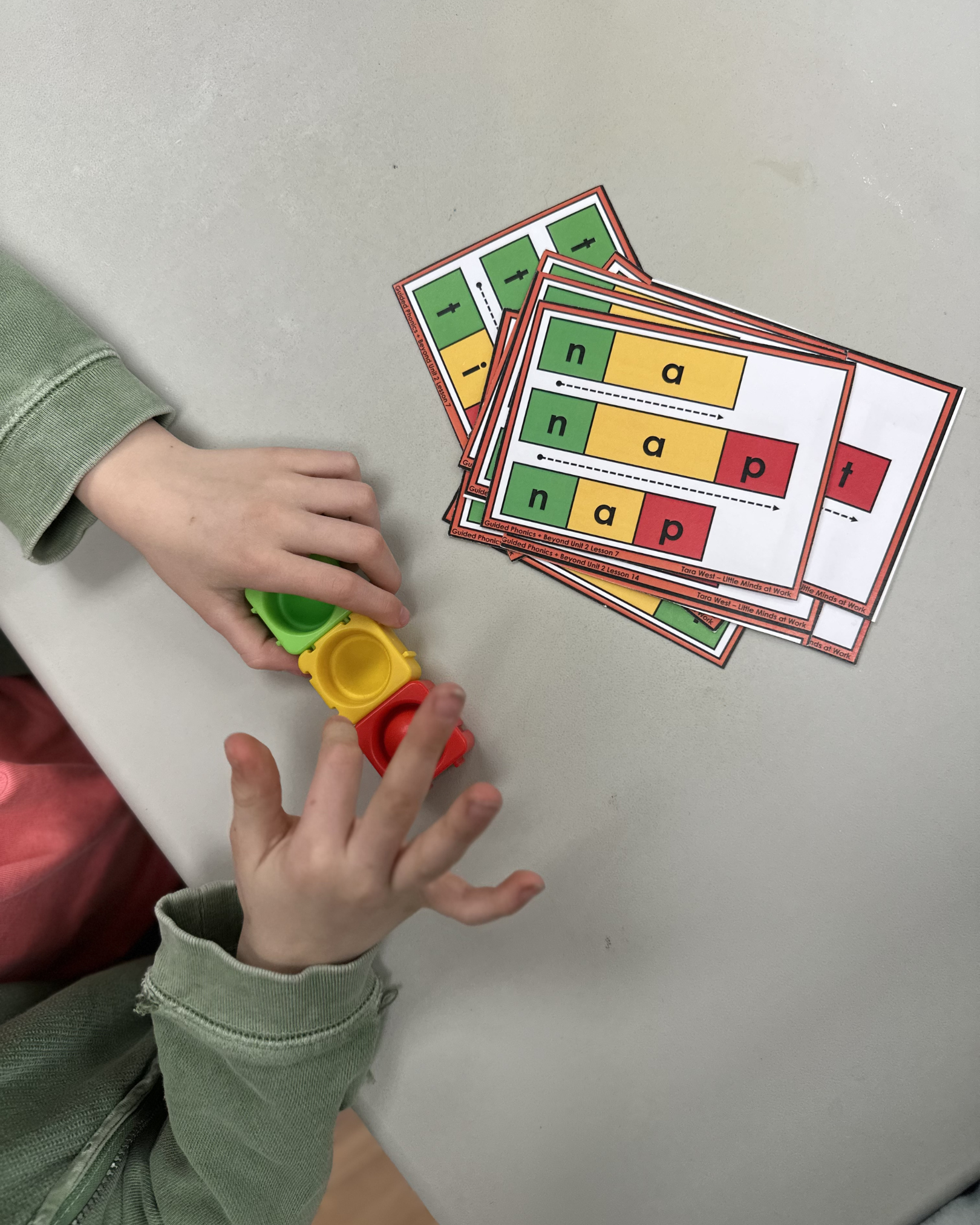 Child's hand holding a colorful educational card game  with letters like 't', 'i', 'n', 'a', 'p' and various dashed lines and arrows for phonics or reading practice.