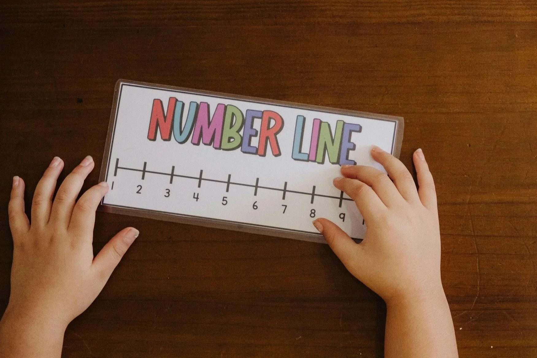 Child's hands holding a number line poster with colorful letters spelling 'Number Line' above a numbered scale from 1 to 10 on a wooden surface. Math tutoring session.