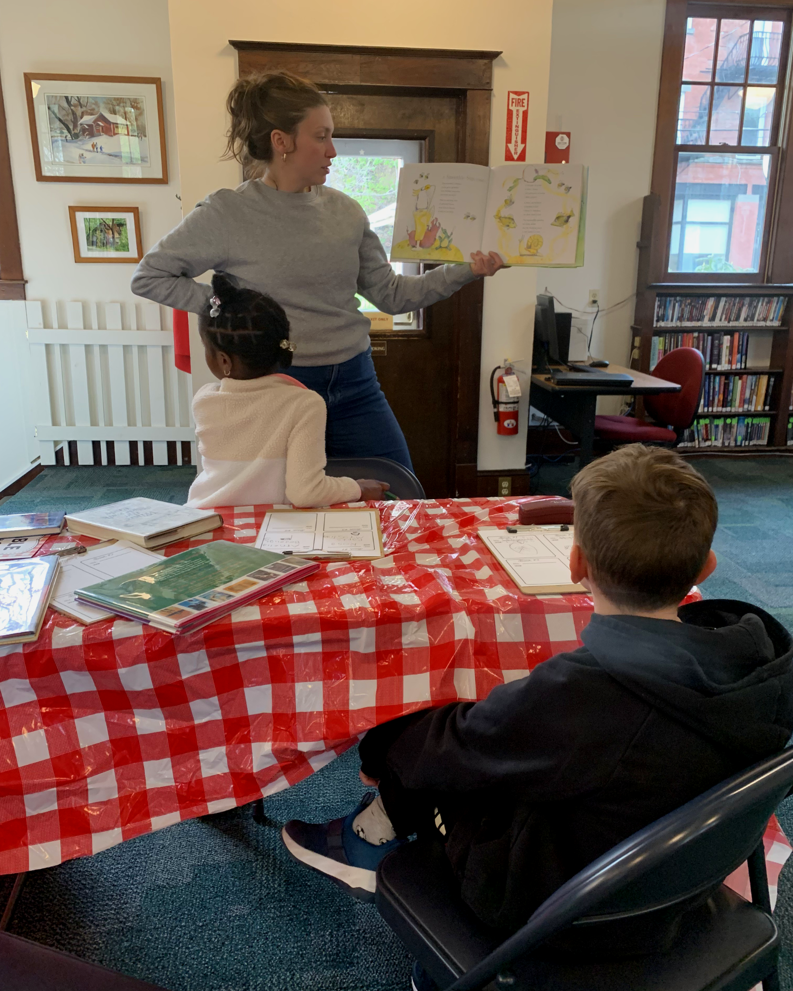 A woman is standing and reading from a children's book to a group of children sitting at a table covered with a red and white checkered tablecloth. The children are listening attentively. The setting appears to be a library or community room with pictures on the wall, a window, a bookshelf filled with books, and a computer desk.
