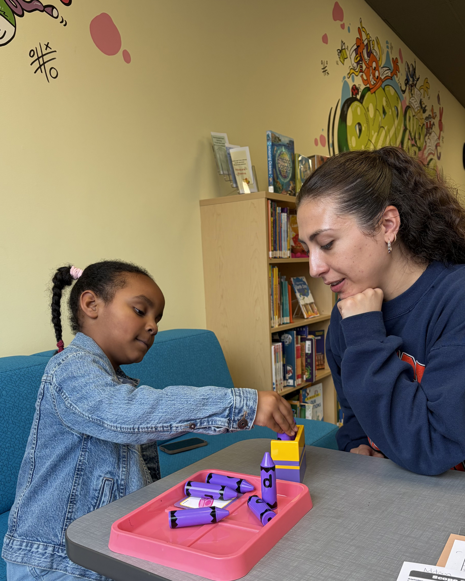 A young girl with braided hair and a woman with earrings and dark hair are engaged in a tabletop activity involving purple and black crayons. They are sitting at a gray table with a pink tray holding crayon pieces. A bookshelf with children's books is behind them in a room with a cartoon mural on the wall.