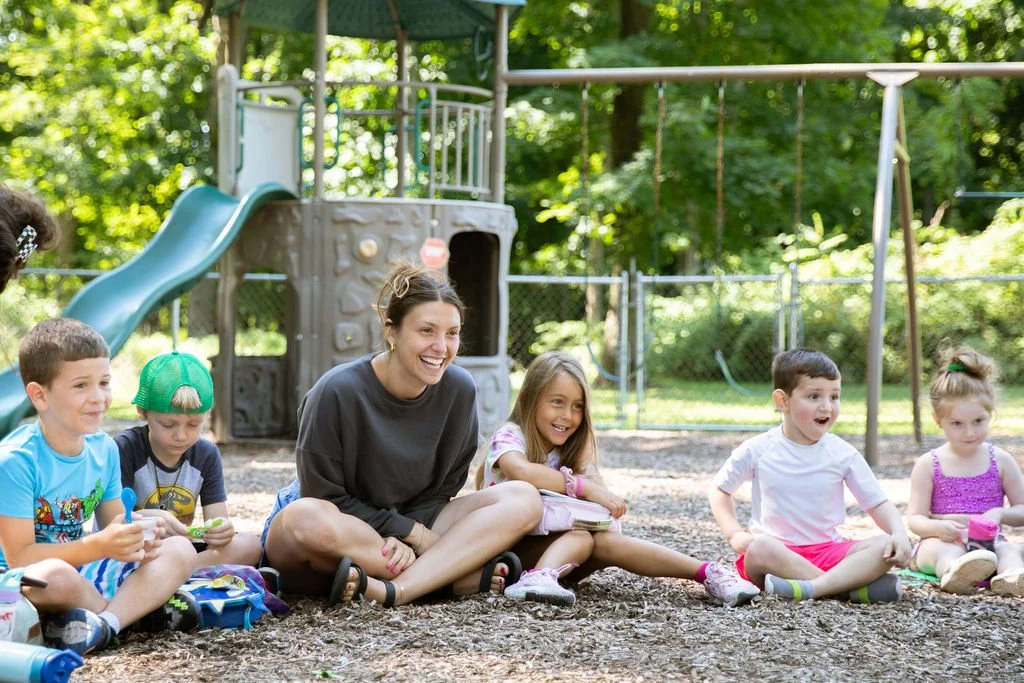 A group of children and a woman sitting on the ground in a playground, smiling and laughing, with a slide and playground equipment in the background.