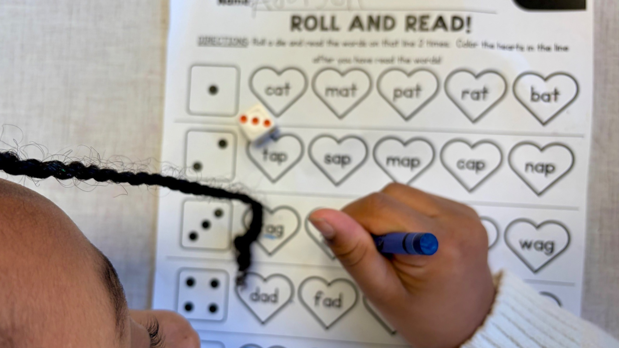 Person working on a homework sheet with heart-shaped words and dice, with a piece of black rope and a blue marker in hand.
