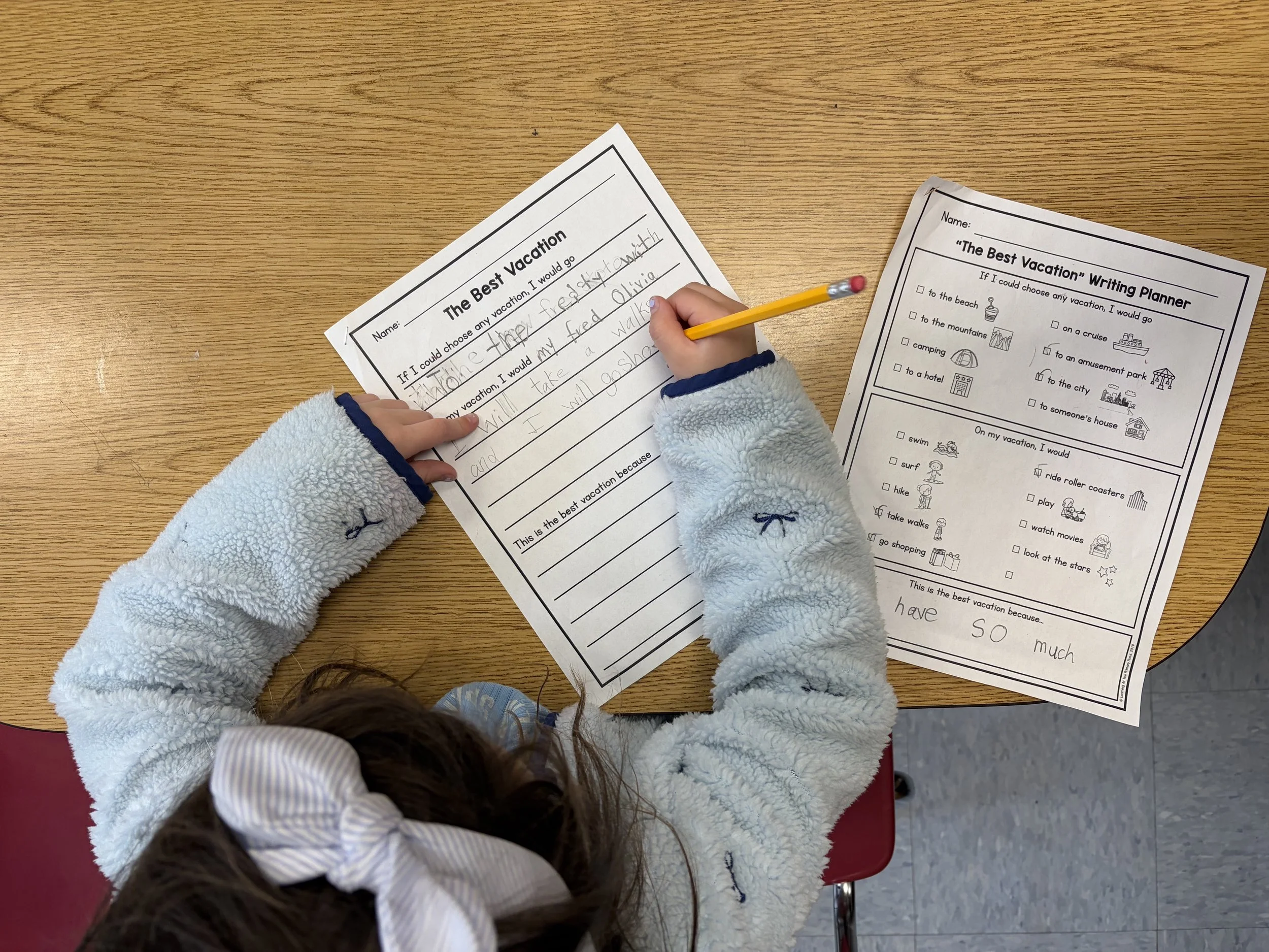 Child writing on a school worksheet titled 'The Best Vacation' on a wooden table, with additional worksheet nearby featuring vacation options with icons. Writing tutoring session.
