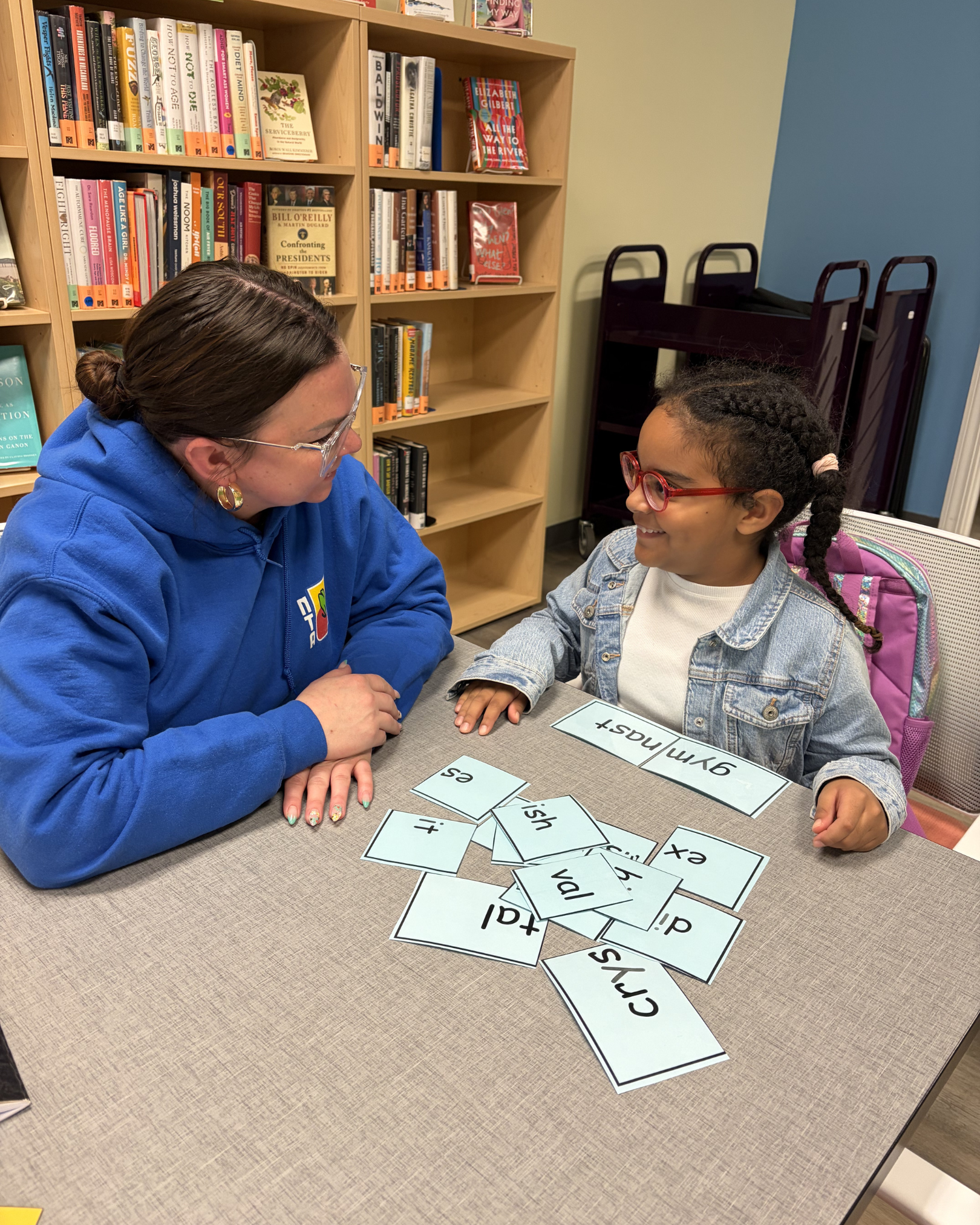 A woman and a girl sitting at a table with alphabet flashcards scattered on it. The woman is wearing a blue hoodie and glasses, smiling while looking at the girl. The girl, wearing red glasses and a denim jacket, is smiling back. In the background, there is a bookshelf filled with books and a cart.