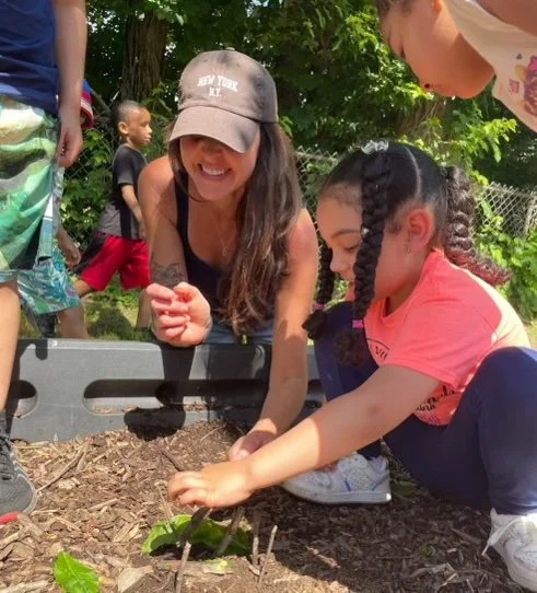 Group of children and an adult planting or tending to a small plant in a garden.
