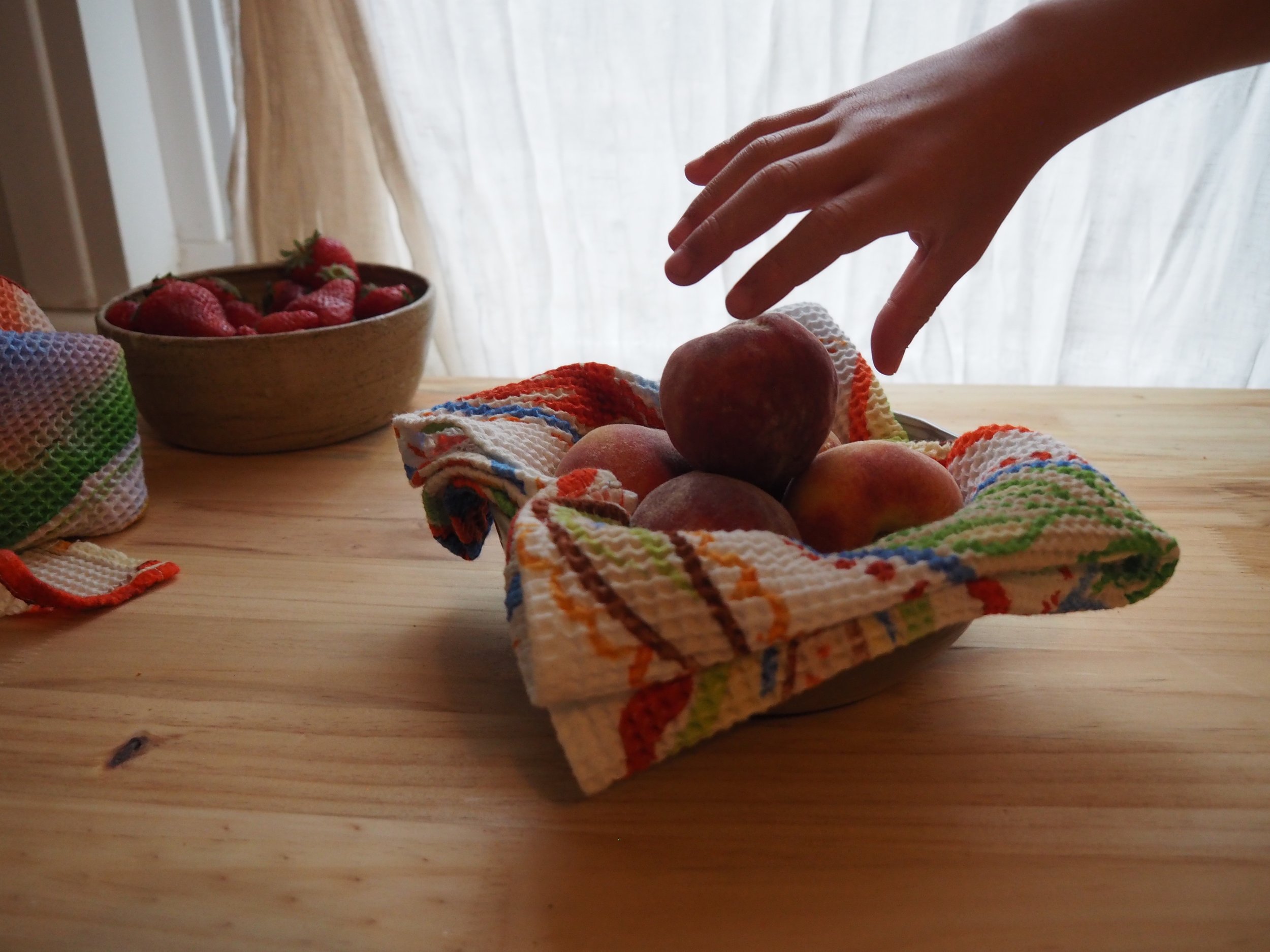 A hand reaching for peaches on a rustic wooden table, with a bowl of strawberries in the background.