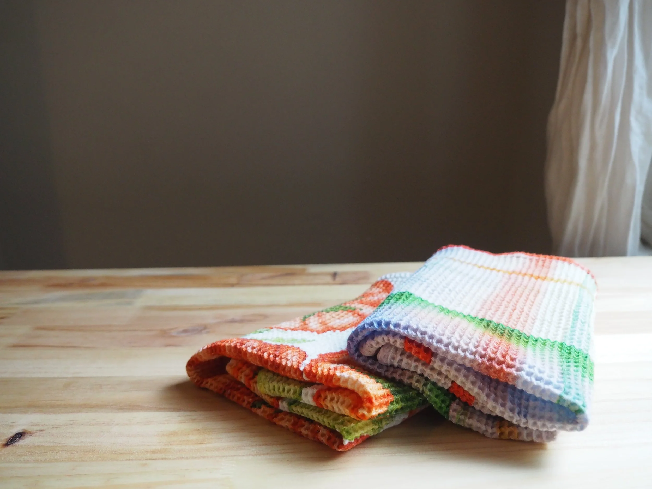 A neatly folded stack of colorful, checkered washcloths on a wooden surface, with a dark wall and a curtain in the background.