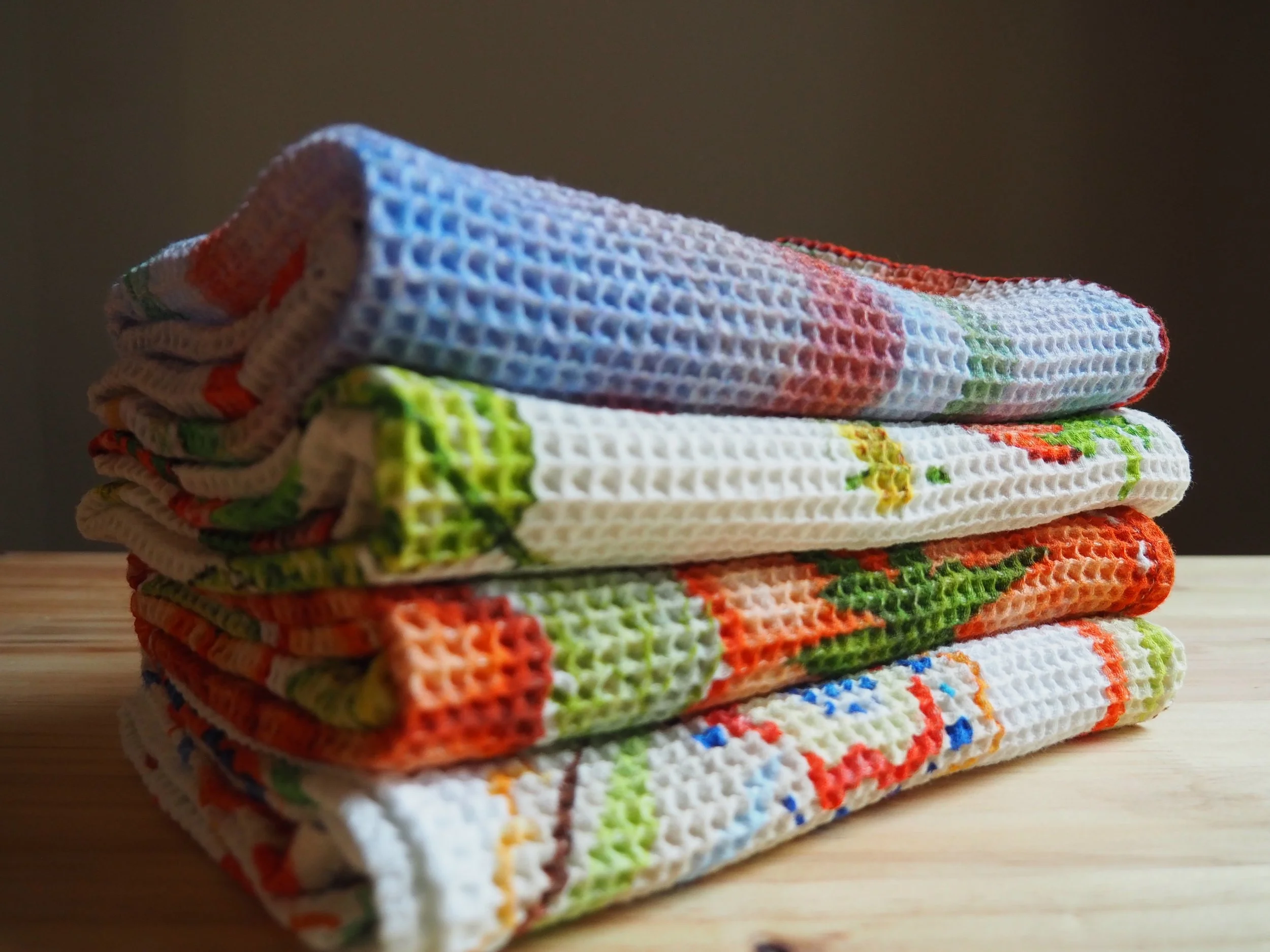 A stack of colorful, hand created patterned tea towels folded and placed on a wooden surface.