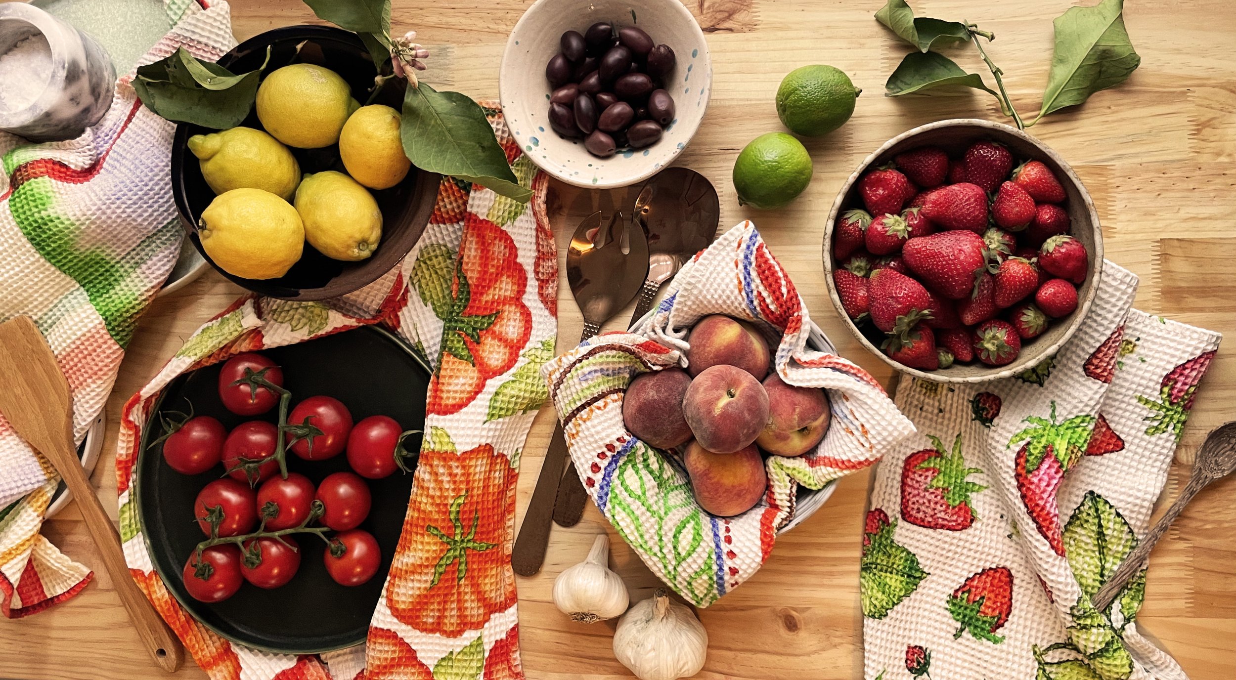 Assorted fresh fruits including lemons, limes, strawberries, peaches, tomatoes, grapes, and black olives, arranged on a wooden table with colorful patterned dish towels.