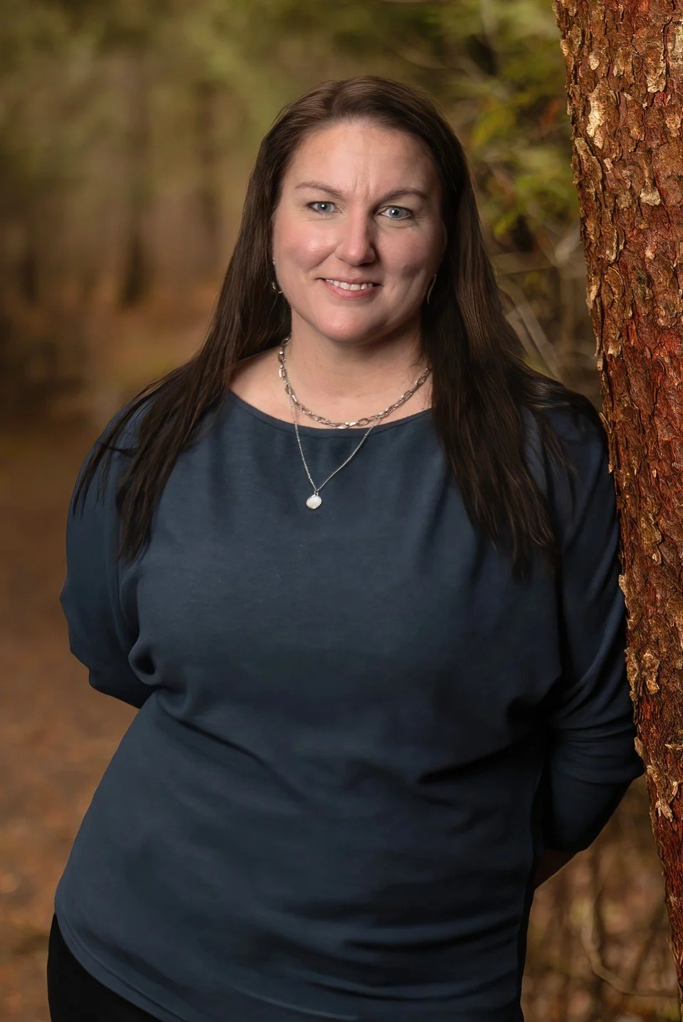 A woman with long dark hair smiling outdoors next to a tree with a blurred forest background.