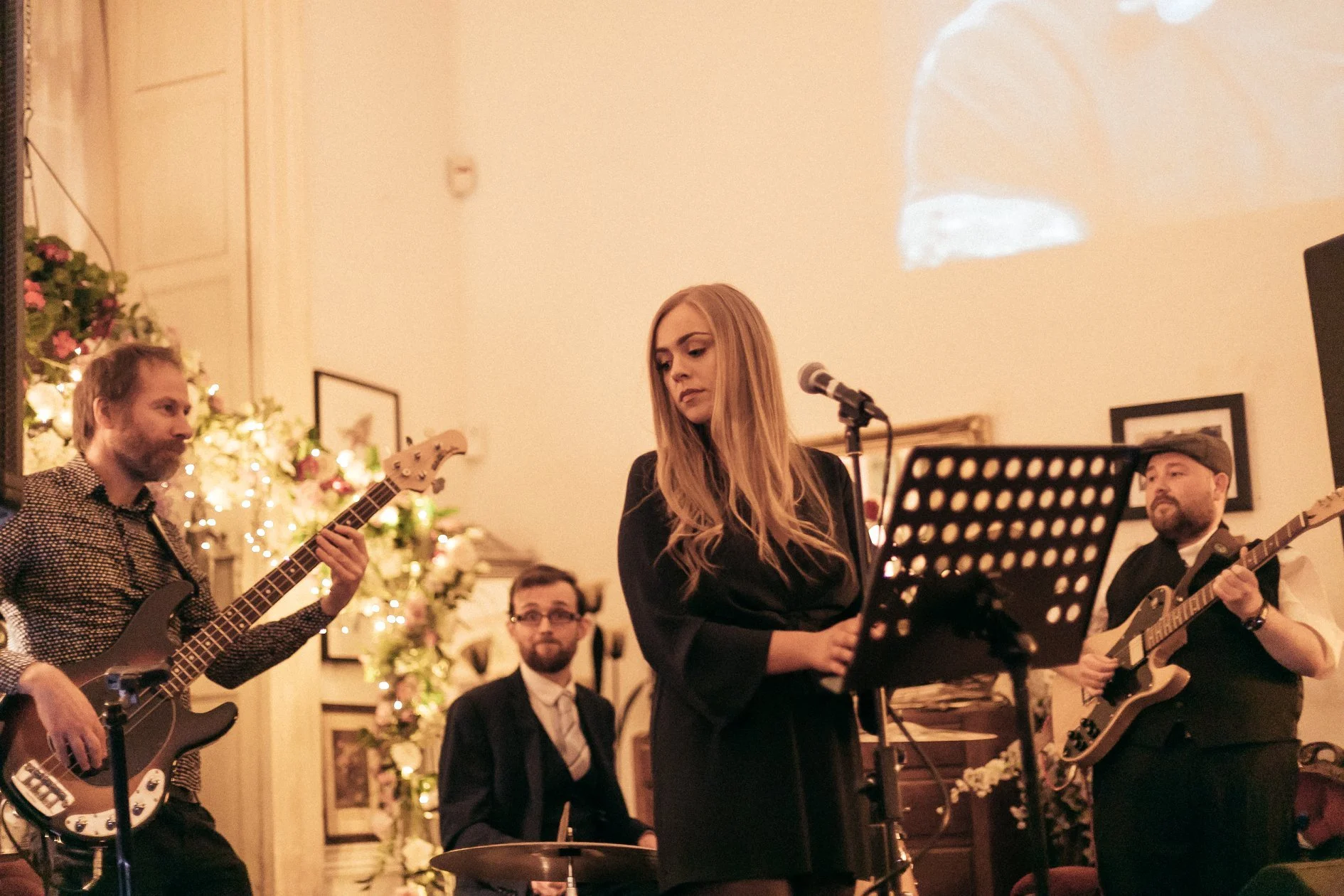 Musicians performing at a cozy indoor event. A woman singing into a microphone, with guitarists and a drummer playing nearby. Warm lighting and floral decorations suggest an intimate concert or gathering.