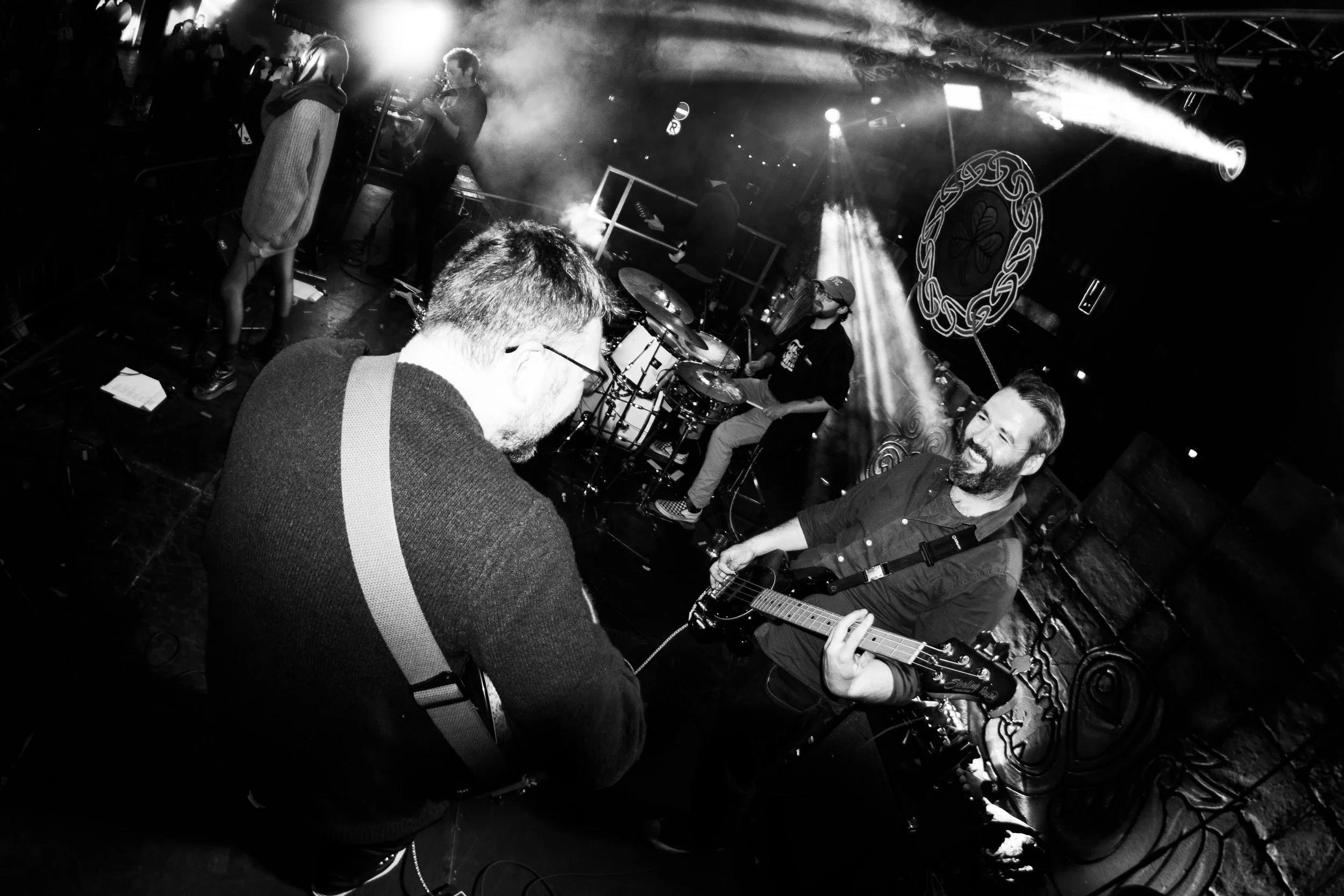 Black and white photo of musicians performing on stage. One person plays drums, another plays guitar, and a woman is standing with her back to the camera. There are stage lights and decorative elements in the background.