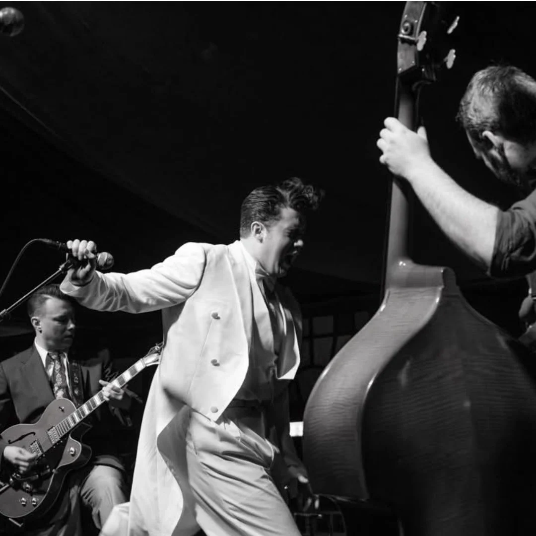 A black and white photo of Elvis Presley performing passionately on stage with a microphone, accompanied by two band members, one playing guitar and the other at a piano.
