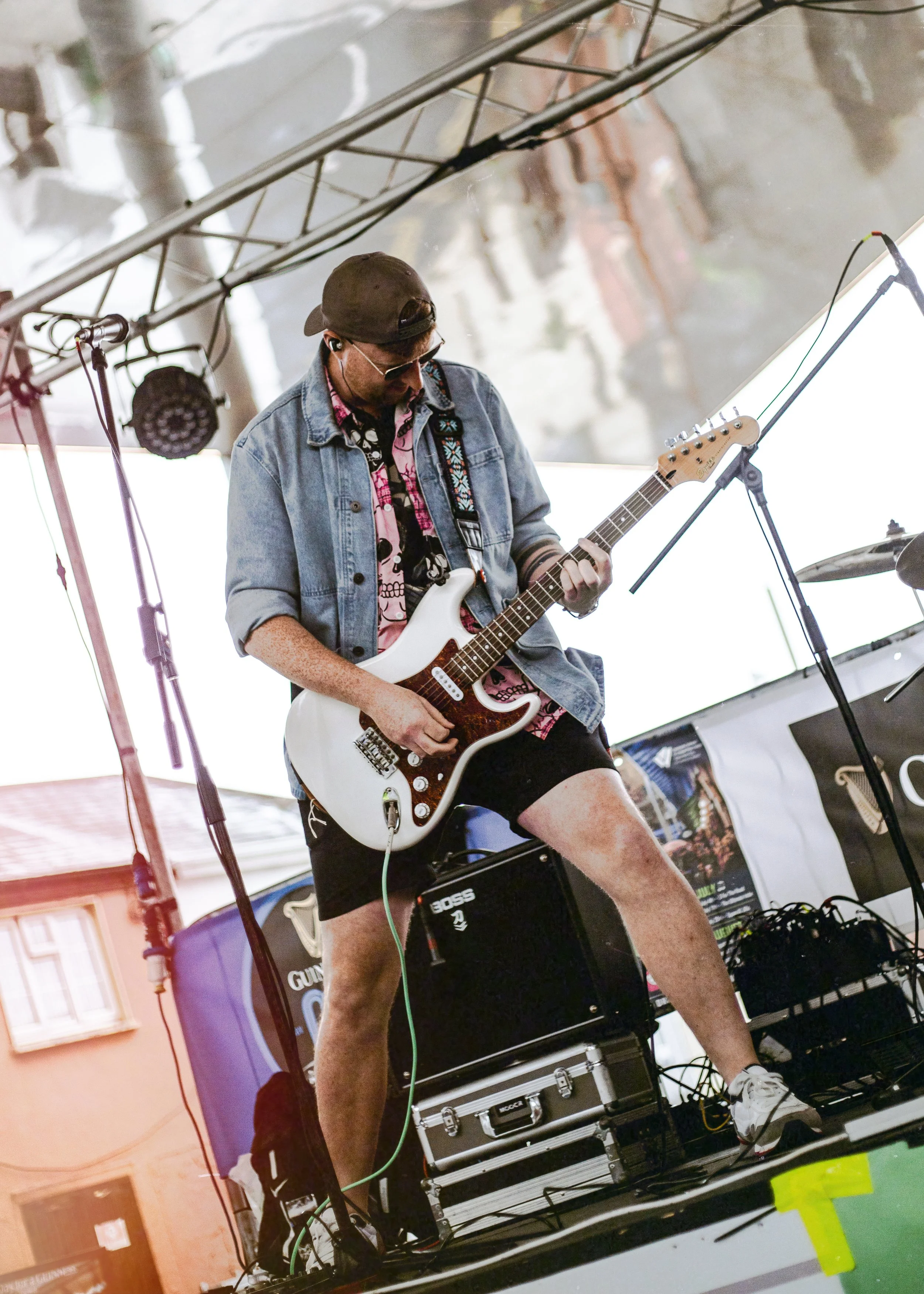 A musician wearing sunglasses, a backwards cap, a denim jacket, and shorts is playing an electric guitar on an outdoor stage, with equipment and sound system surrounding him.