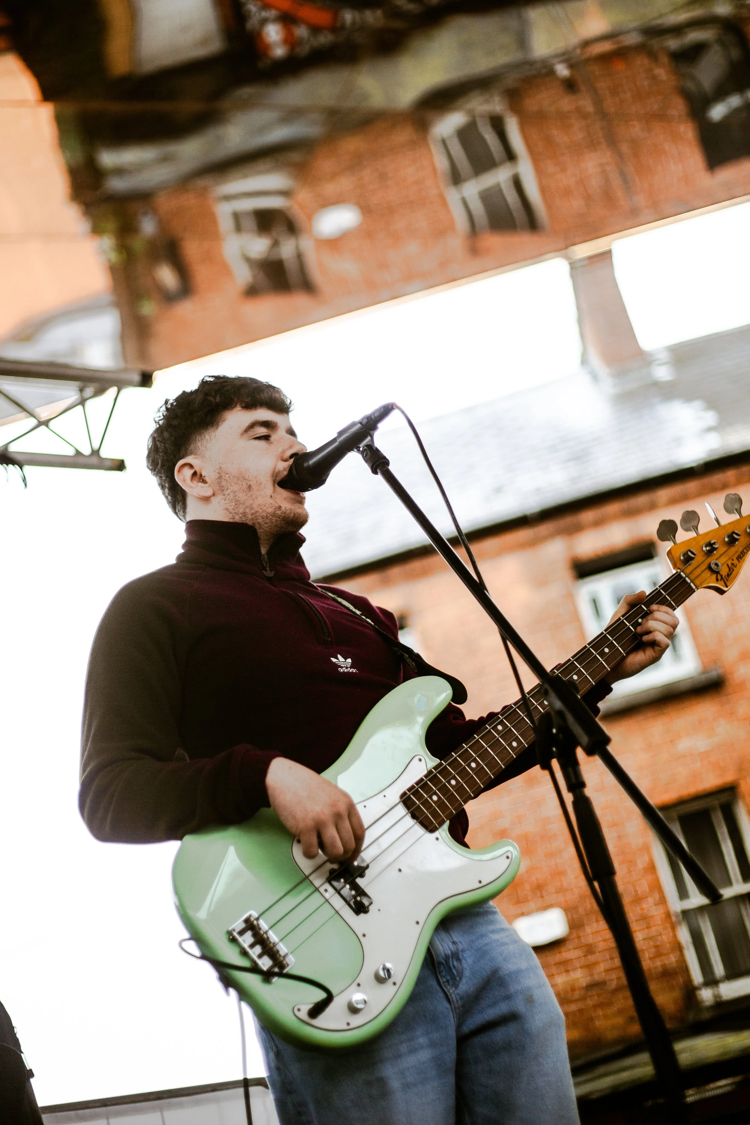 A young man singing into a microphone and playing a light green electric bass guitar during an outdoor performance, with a brick building and overcast sky in the background.
