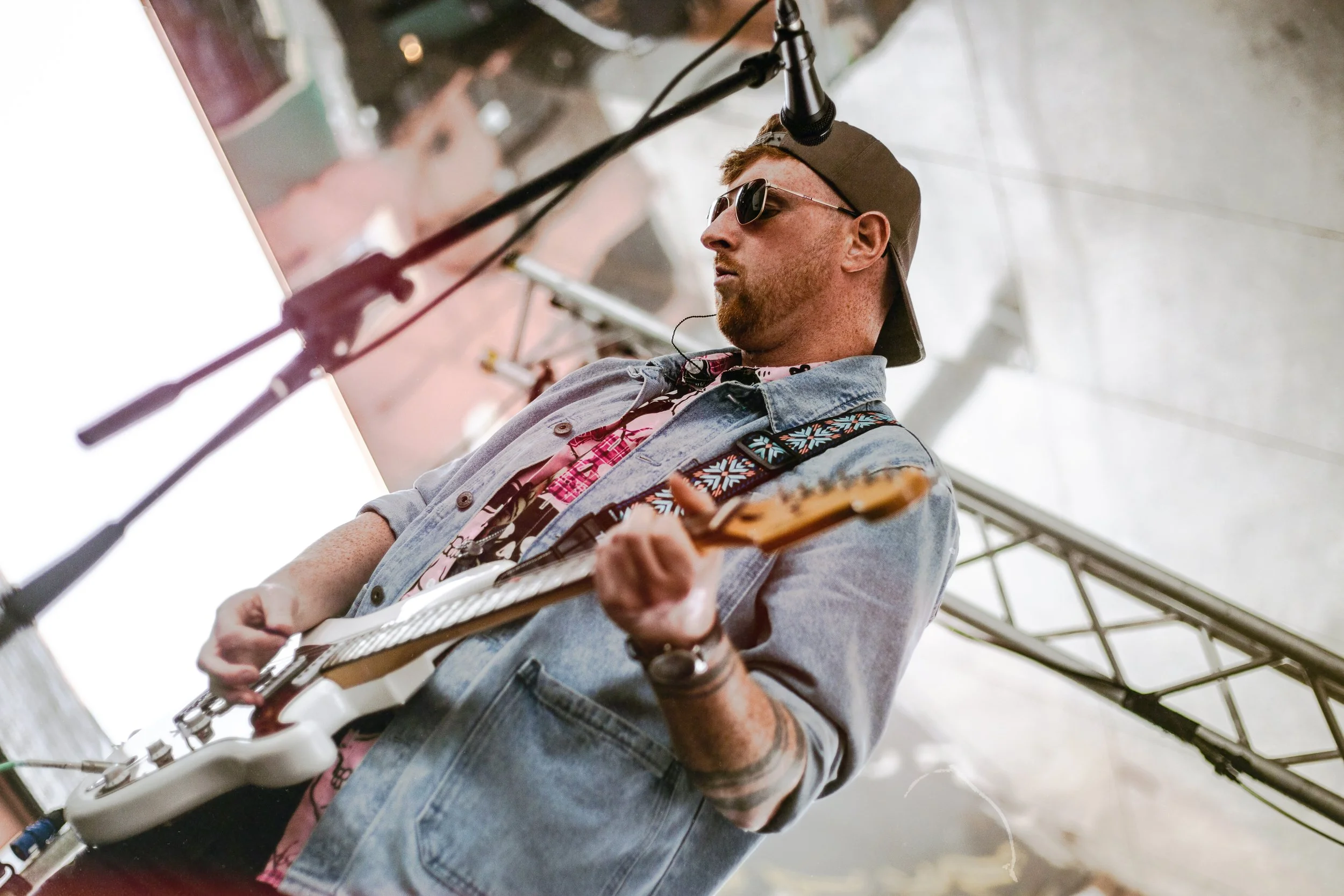 A man wearing sunglasses, a baseball cap backward, a denim jacket, and a pink patterned shirt playing an electric guitar on stage under a tent.